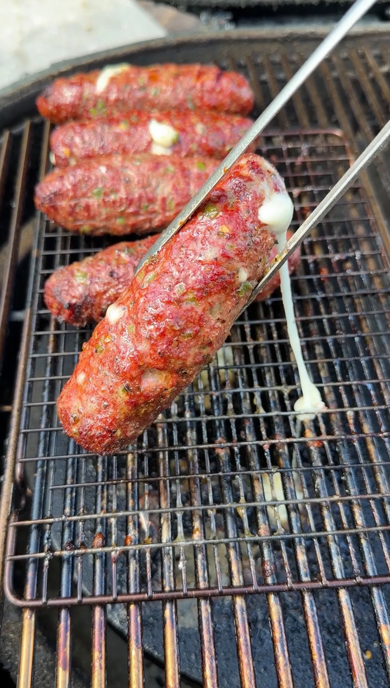 Cheese stuffed burger dogs on a charcoal grill, with melted cheese oozing out, held by metal tongs