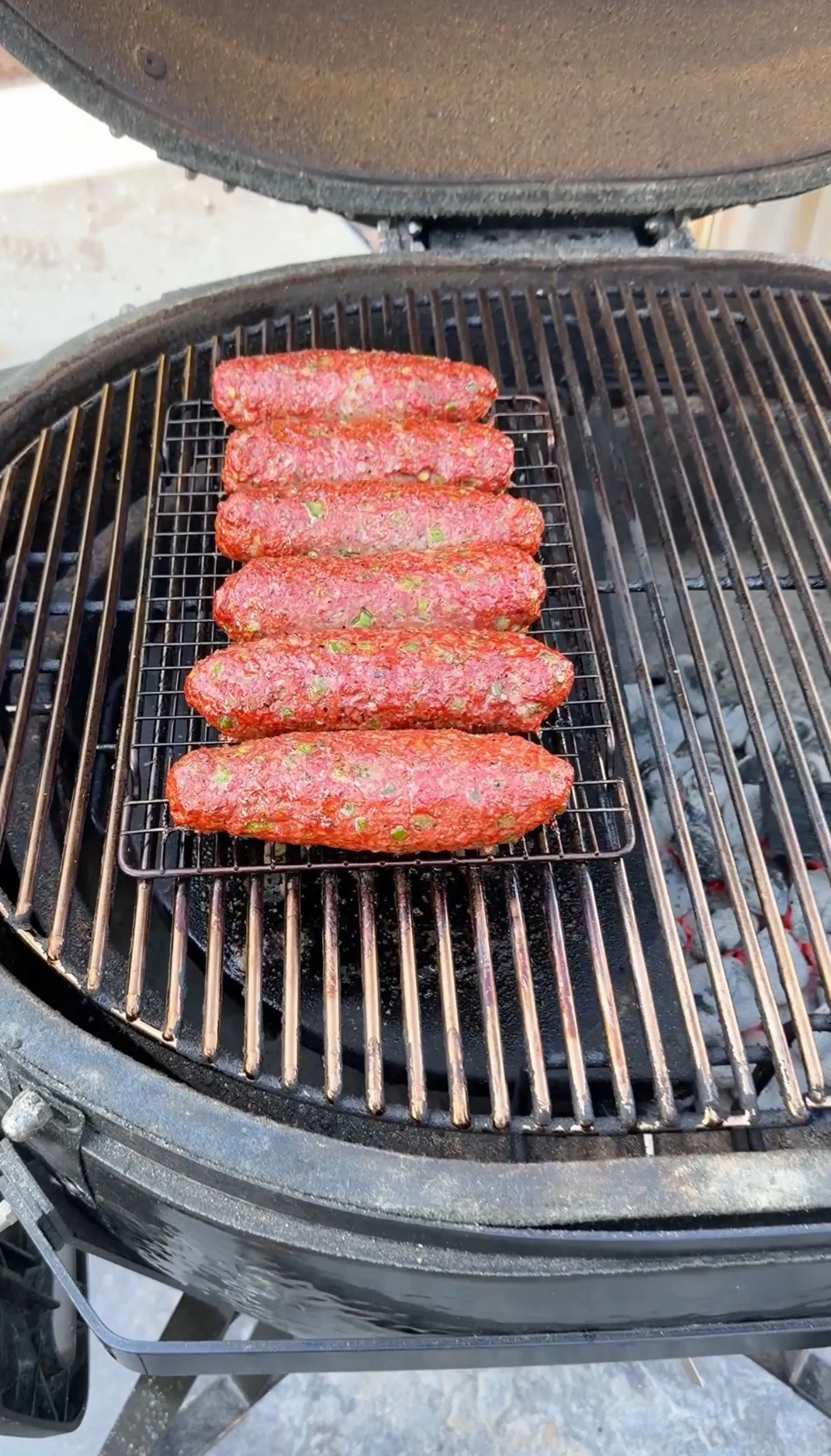 Six raw cheese stuffed burger dogs on a wire rack over hot charcoal in a kamado grill