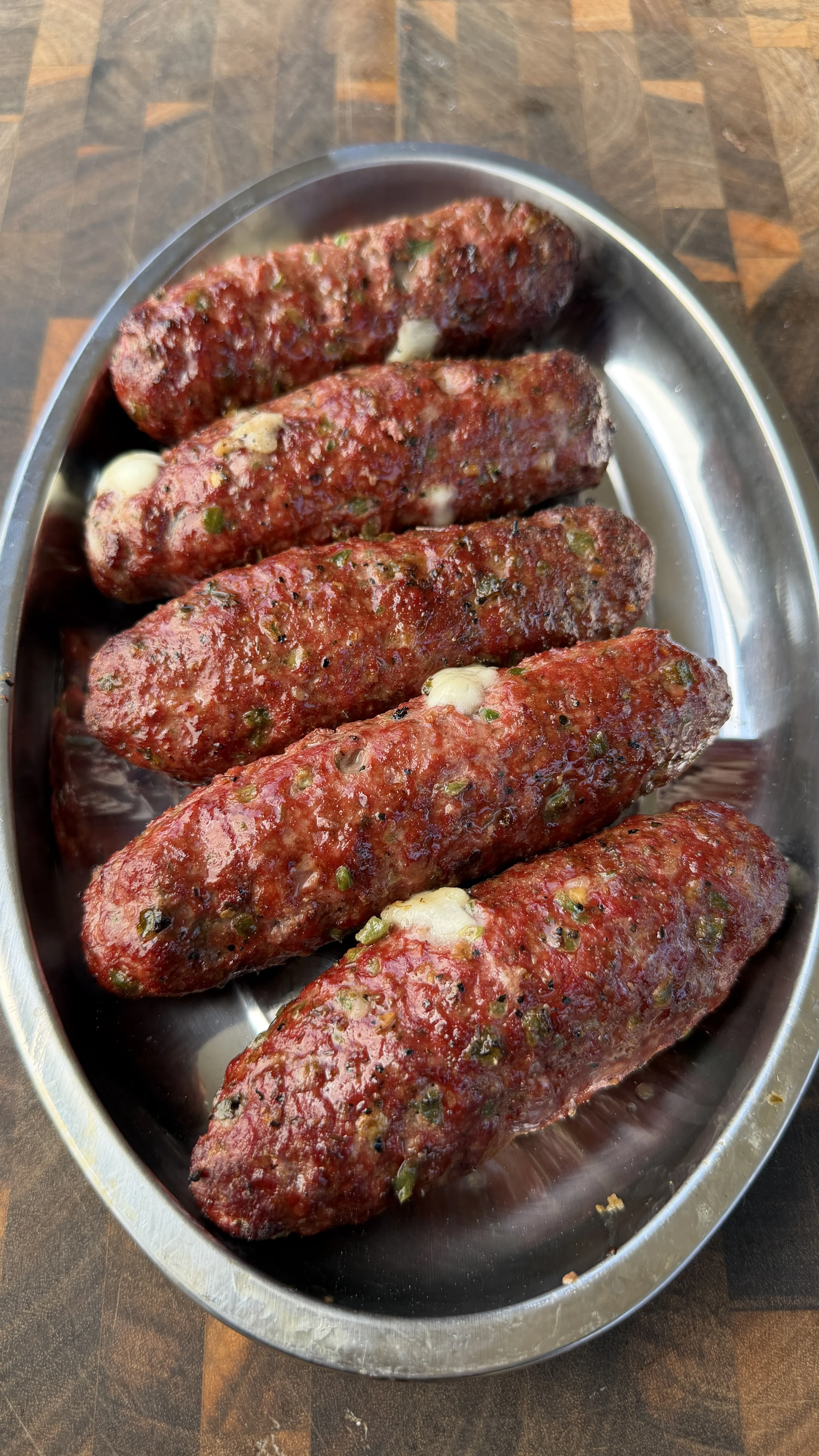 Six juicy cheese-stuffed burger dogs arranged in an oval stainless steel serving dish on a wooden cutting board