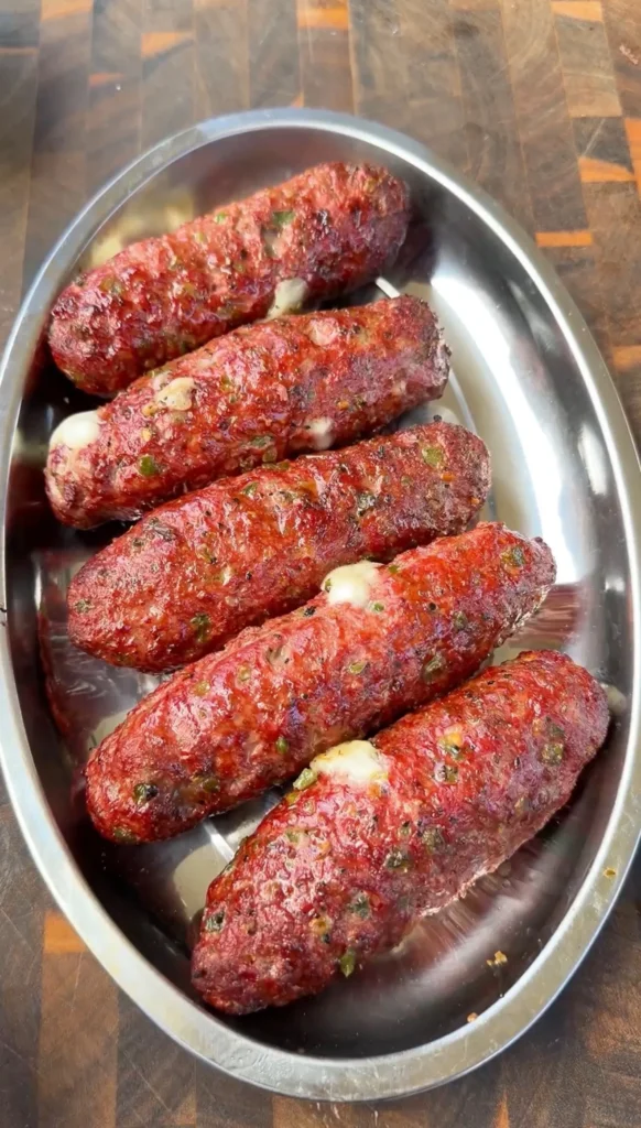 Six grilled cheese-stuffed burger dogs arranged in an oval stainless steel serving dish on a wooden board