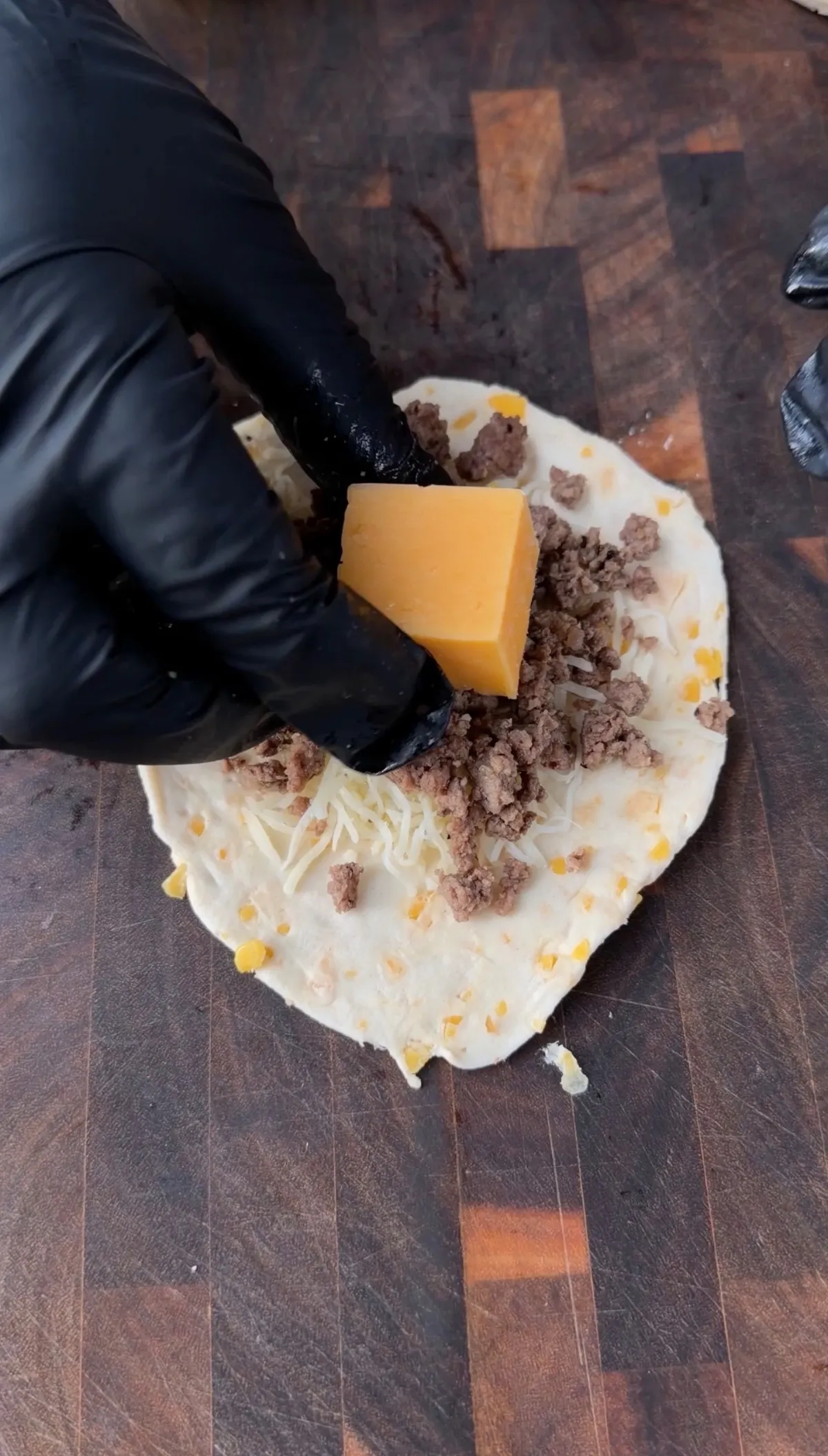 Black-gloved hand placing cheddar cheese block on biscuit dough topped with seasoned ground beef and shredded cheese