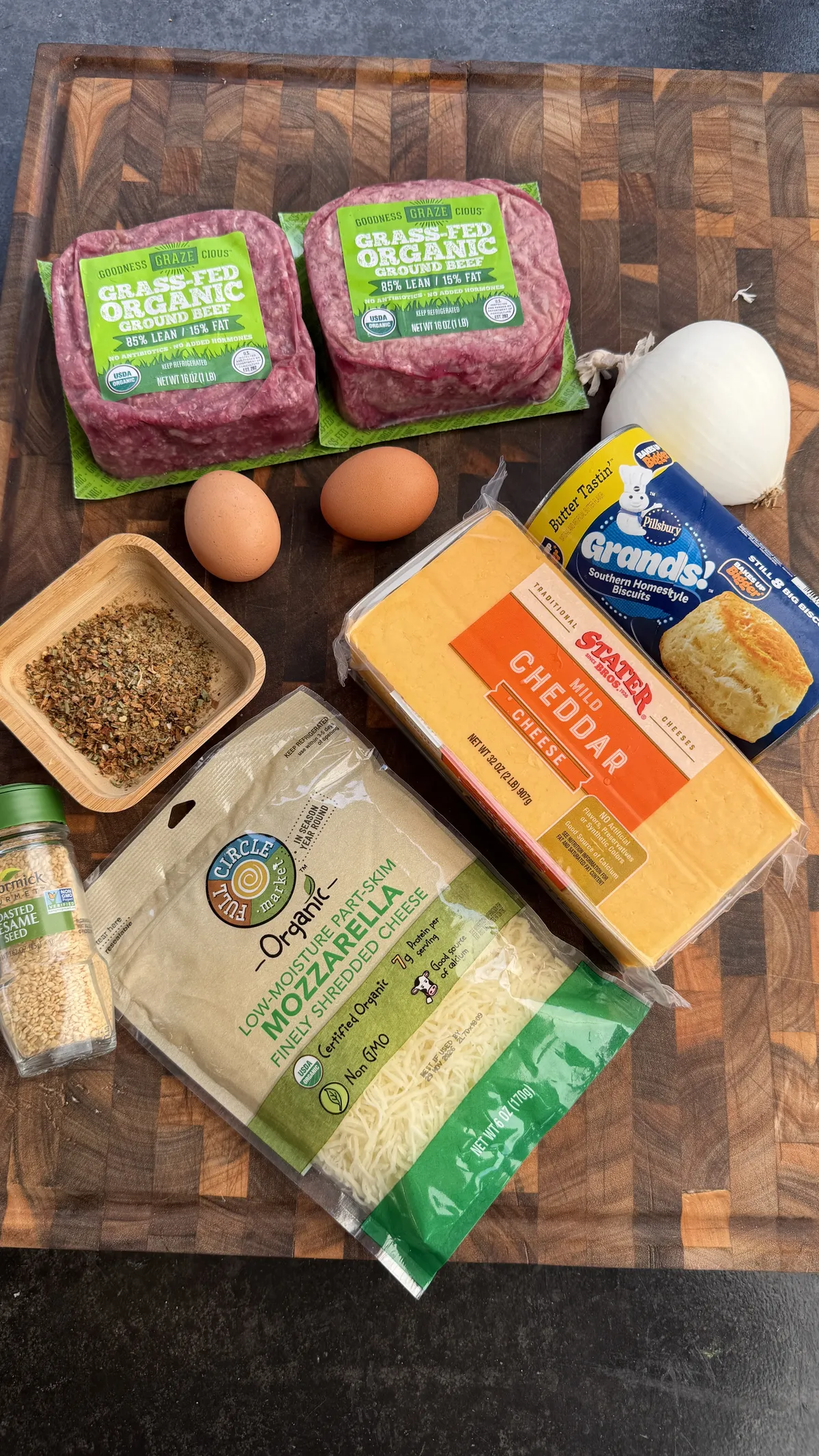 Cheeseburger bomb ingredients on wooden board: ground beef, eggs, cheddar, mozzarella, biscuits, onion, and seasonings