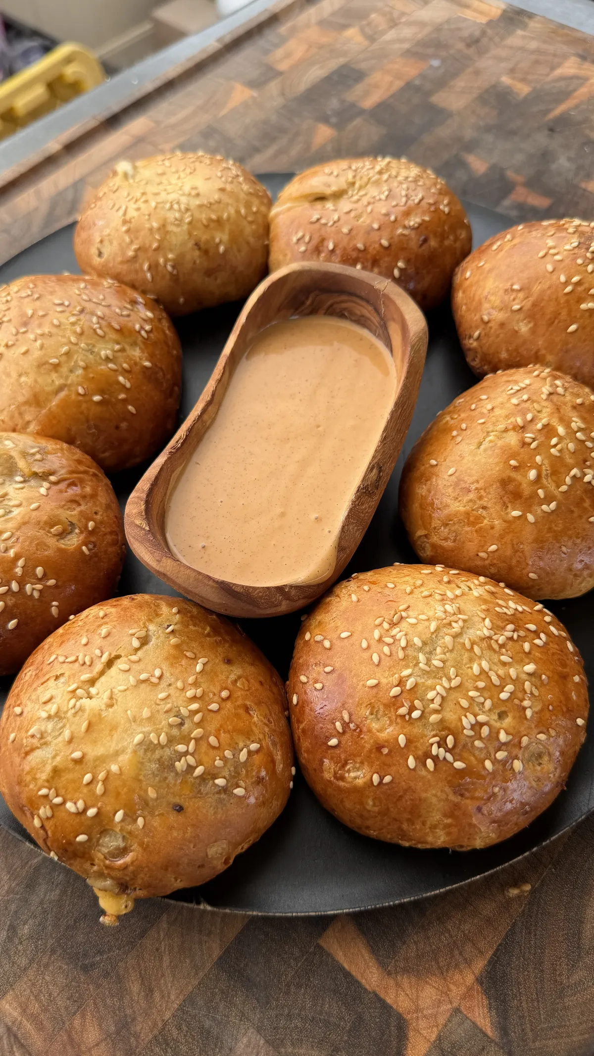 Golden sesame-topped cheeseburger bombs arranged on a dark plate with creamy dipping sauce in a wooden bowl