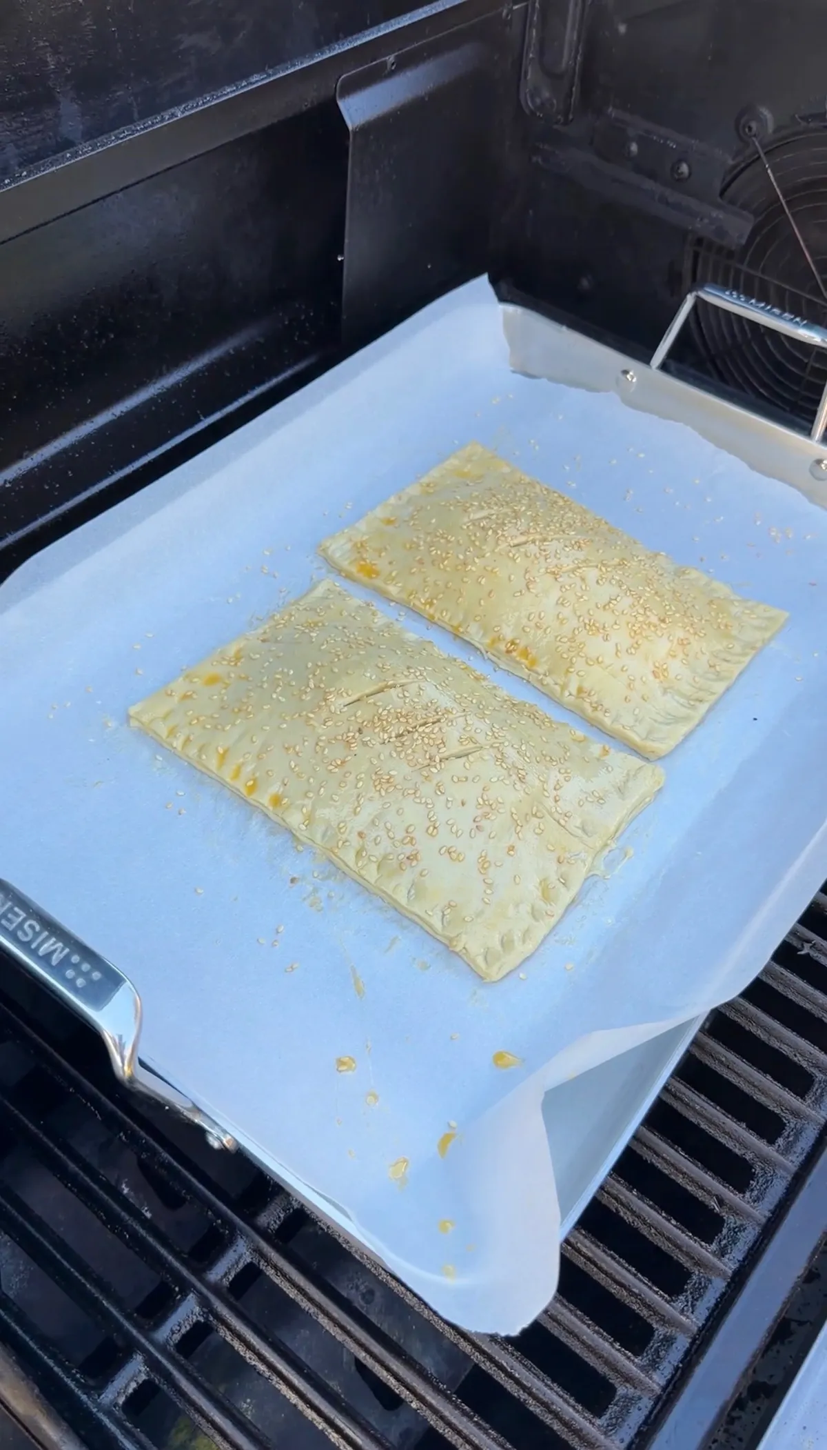 Two unbaked cheeseburger puff pastry hot pockets topped with sesame seeds on parchment-lined baking sheet on a grill
