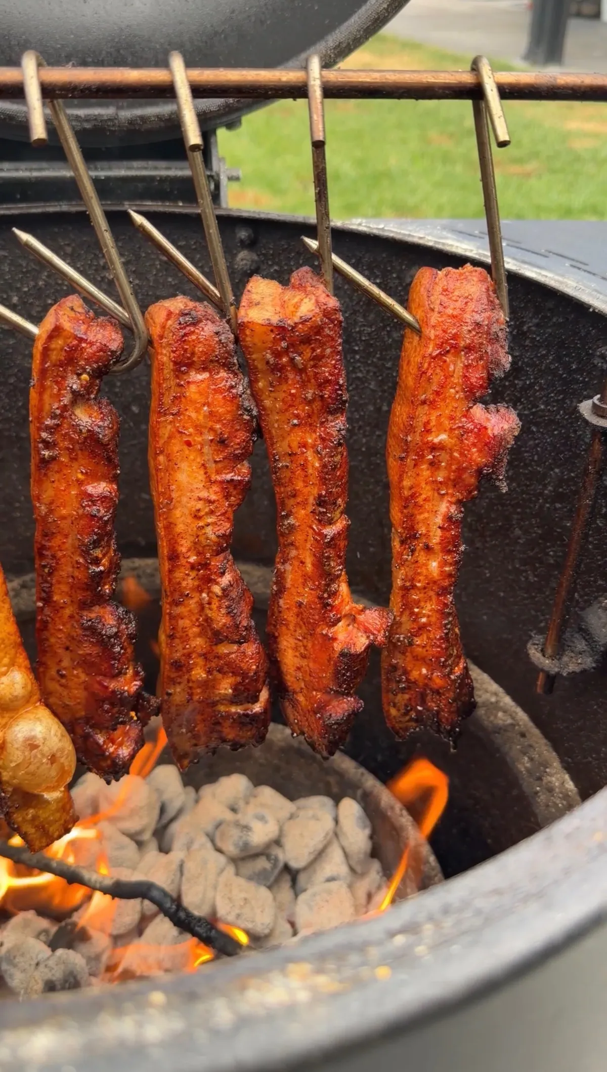 Seasoned pork chicharron strips hanging on hooks over glowing charcoal flames in a round smoker grill