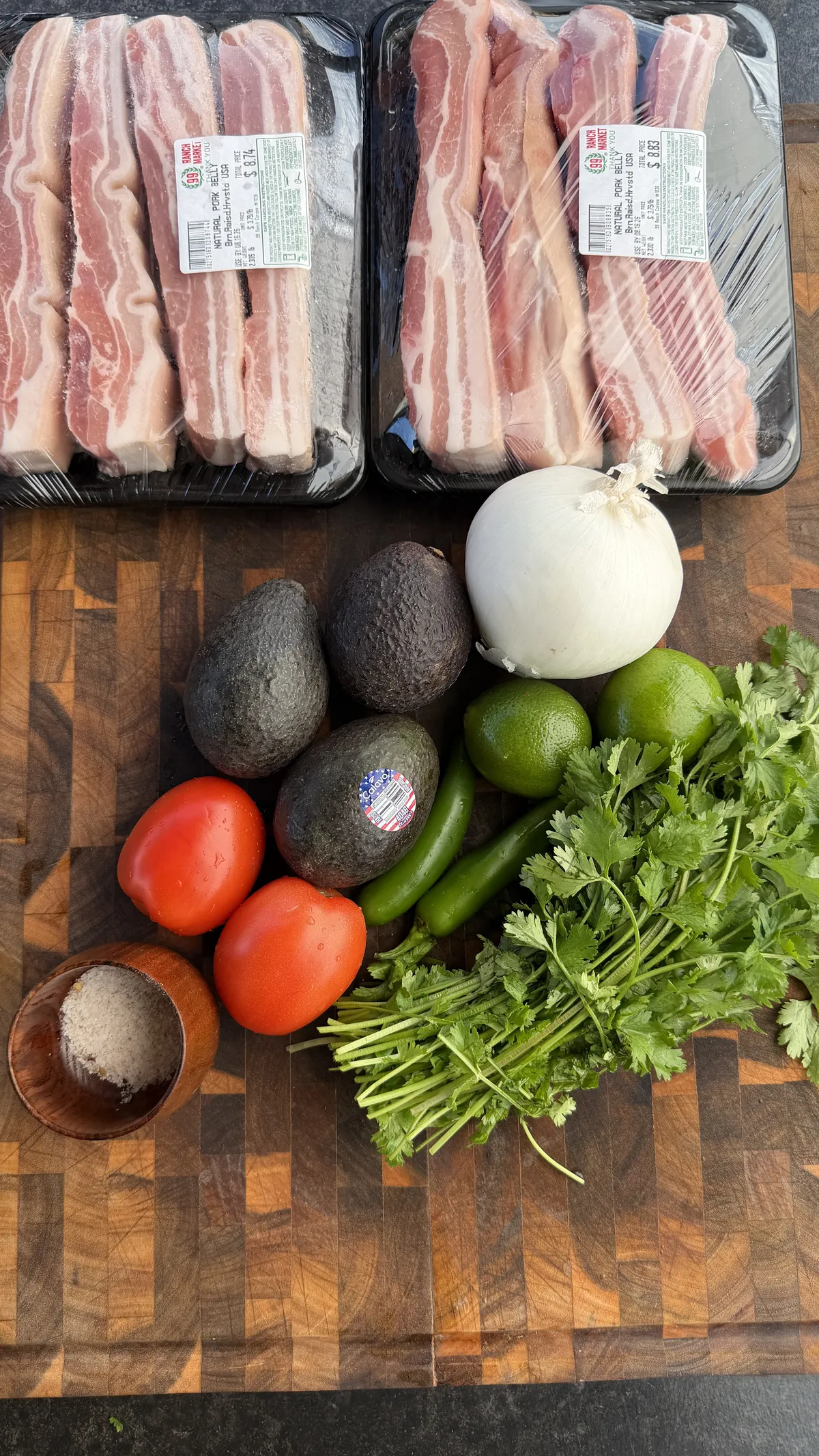 Raw pork belly slabs with avocados, tomatoes, white onion, jalapeños, limes, cilantro, and salt on a wooden cutting board