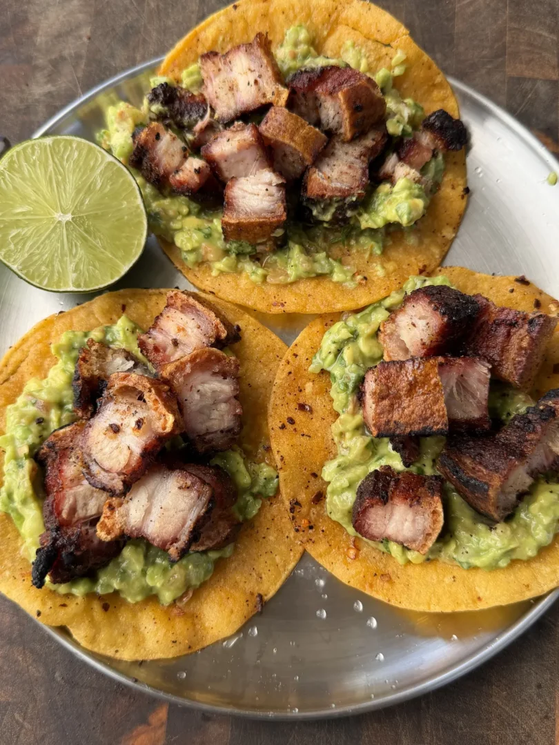 Three crispy chicharron tacos topped with chunky guacamole on corn tortillas, served on a silver plate with lime wedge