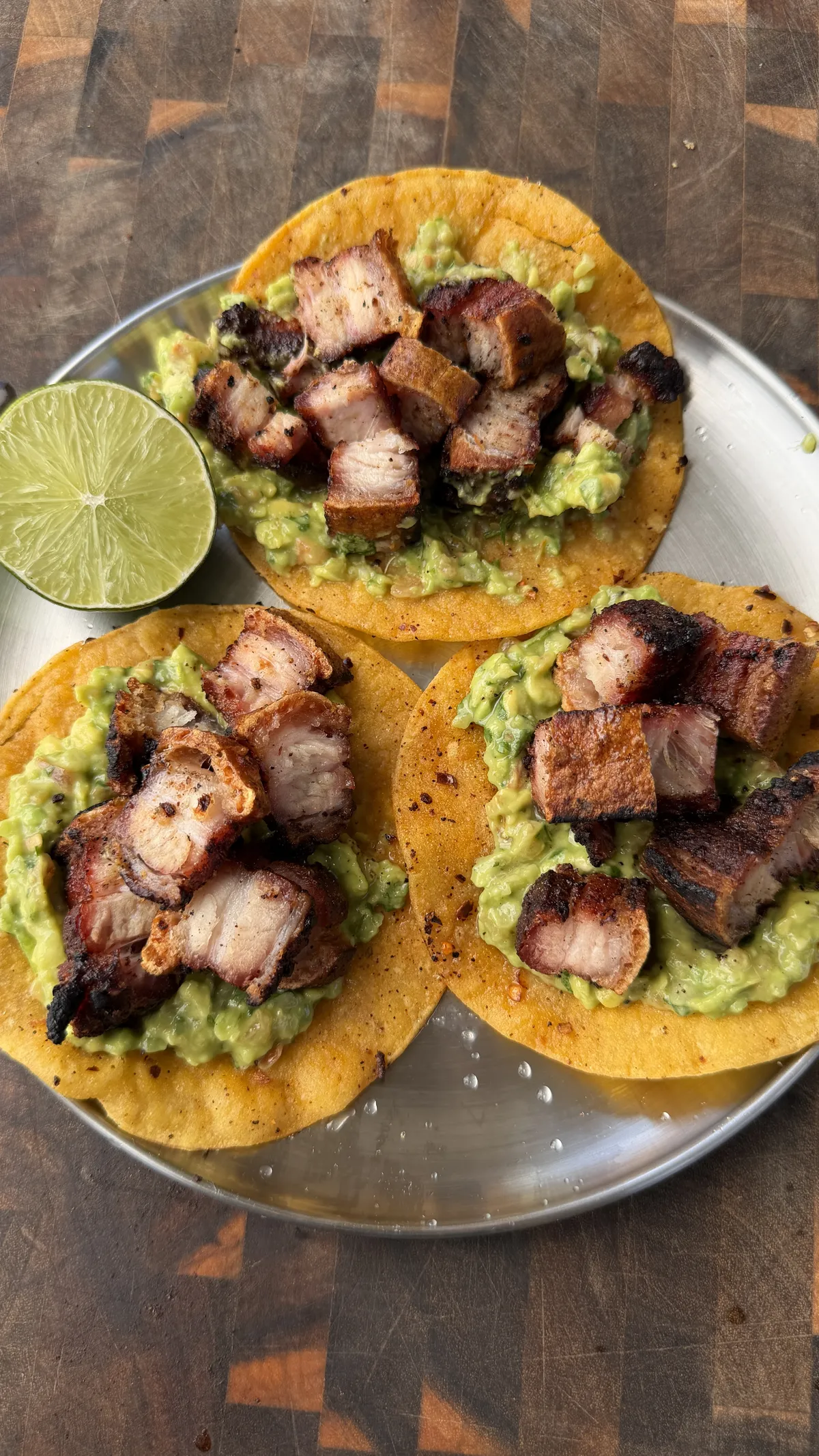 Three crispy chicharron tacos topped with chunky guacamole on corn tortillas, served on a silver plate with lime wedge