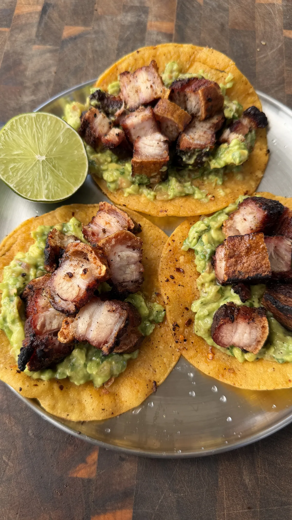 Three crispy chicharron tacos on corn tortillas with guacamole on a silver plate, served with a lime half