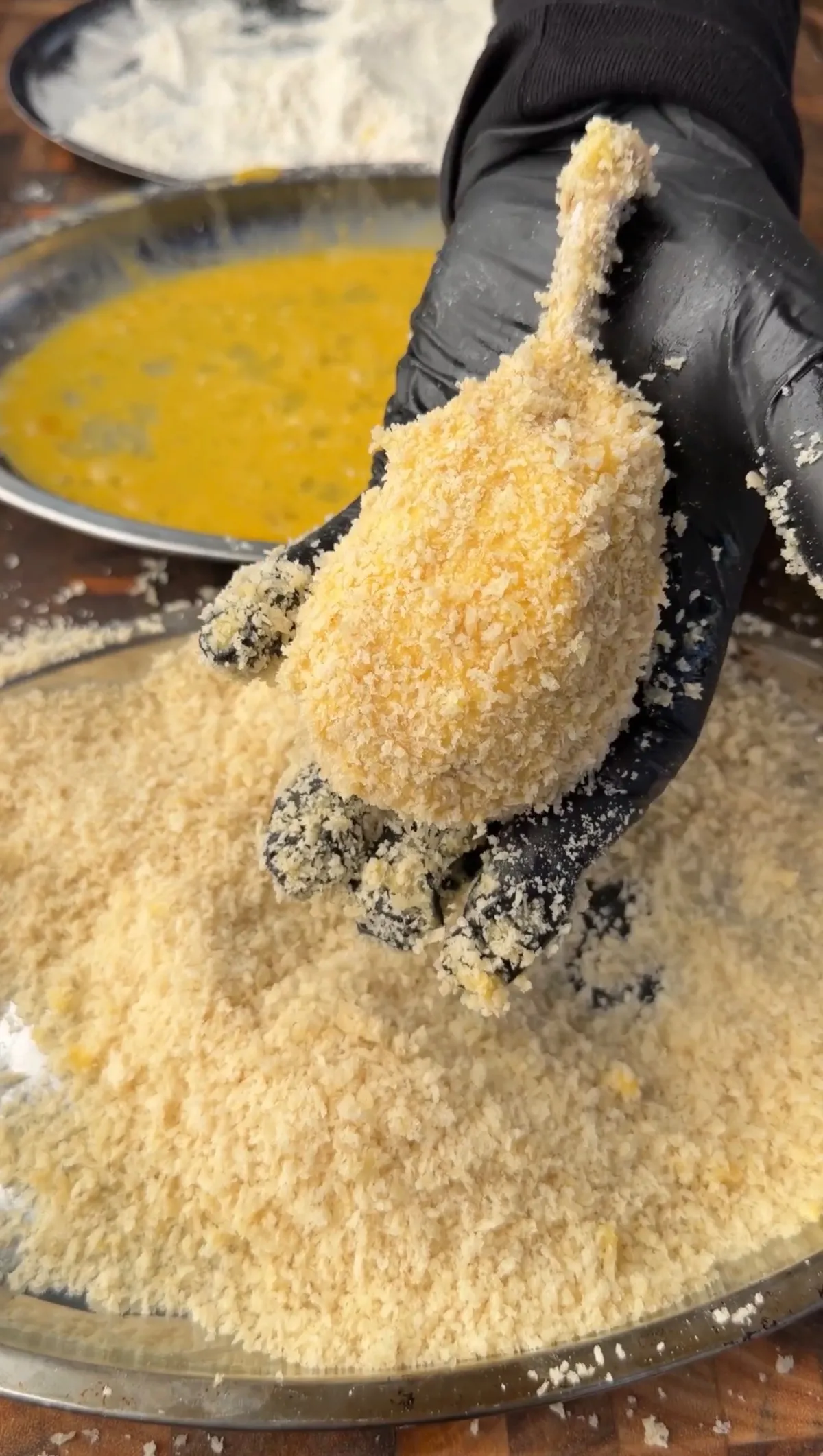 Gloved hand coating a shaped chicken potato cheese bomb in panko breadcrumbs over a metal tray