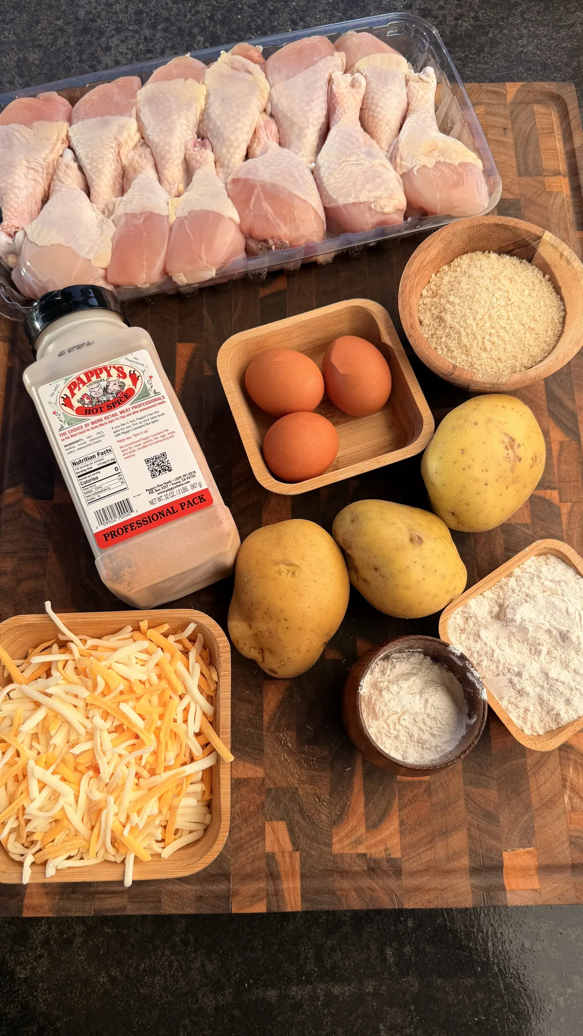 Ingredients for Chicken Potato Cheese Bombs: drumsticks, golden potatoes, shredded cheese, eggs, panko breadcrumbs, flour, and Pappy's Hot Spice seasoning on a wooden cutting board