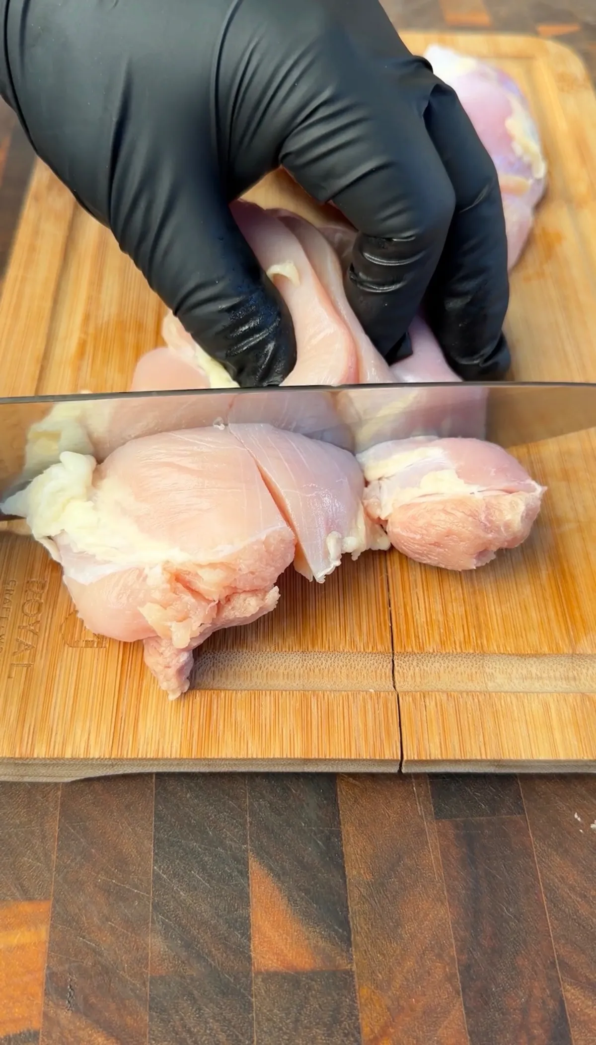 Boneless skinless chicken thighs being cut into 1-inch bite-size pieces on a bamboo cutting board with black gloved hands