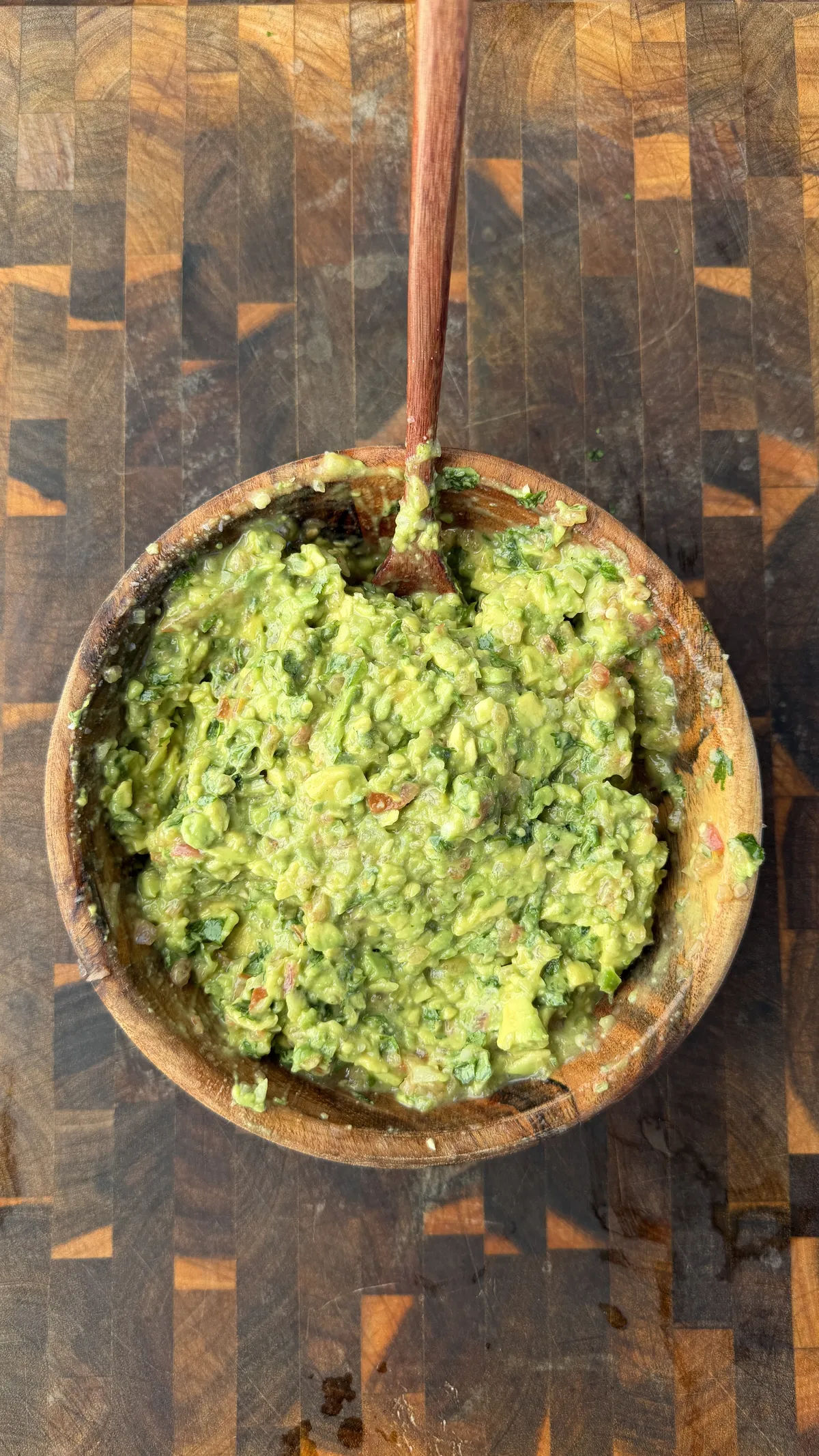 Freshly made chunky guacamole with cilantro in a rustic wooden bowl with a wooden spoon on a butcher block