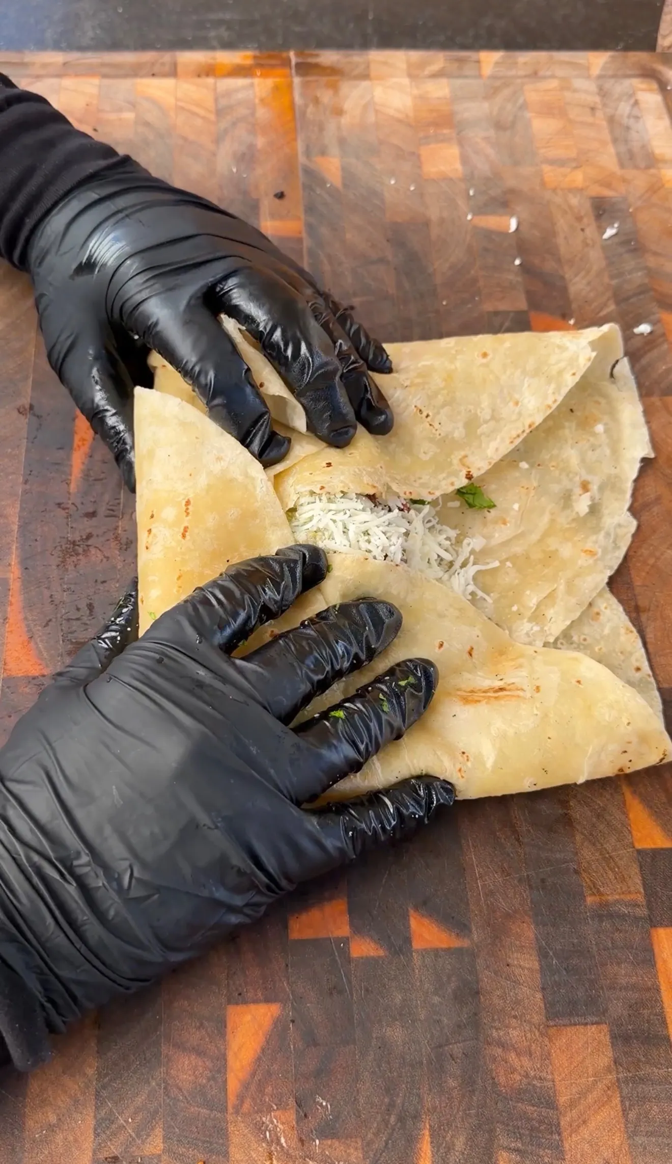 Gloved hands folding a crispy carne asada chimichanga with shredded cheese on a wooden cutting board