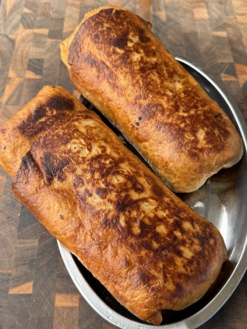 Two golden-brown crispy carne asada chimichangas on a stainless steel plate atop a wooden cutting board