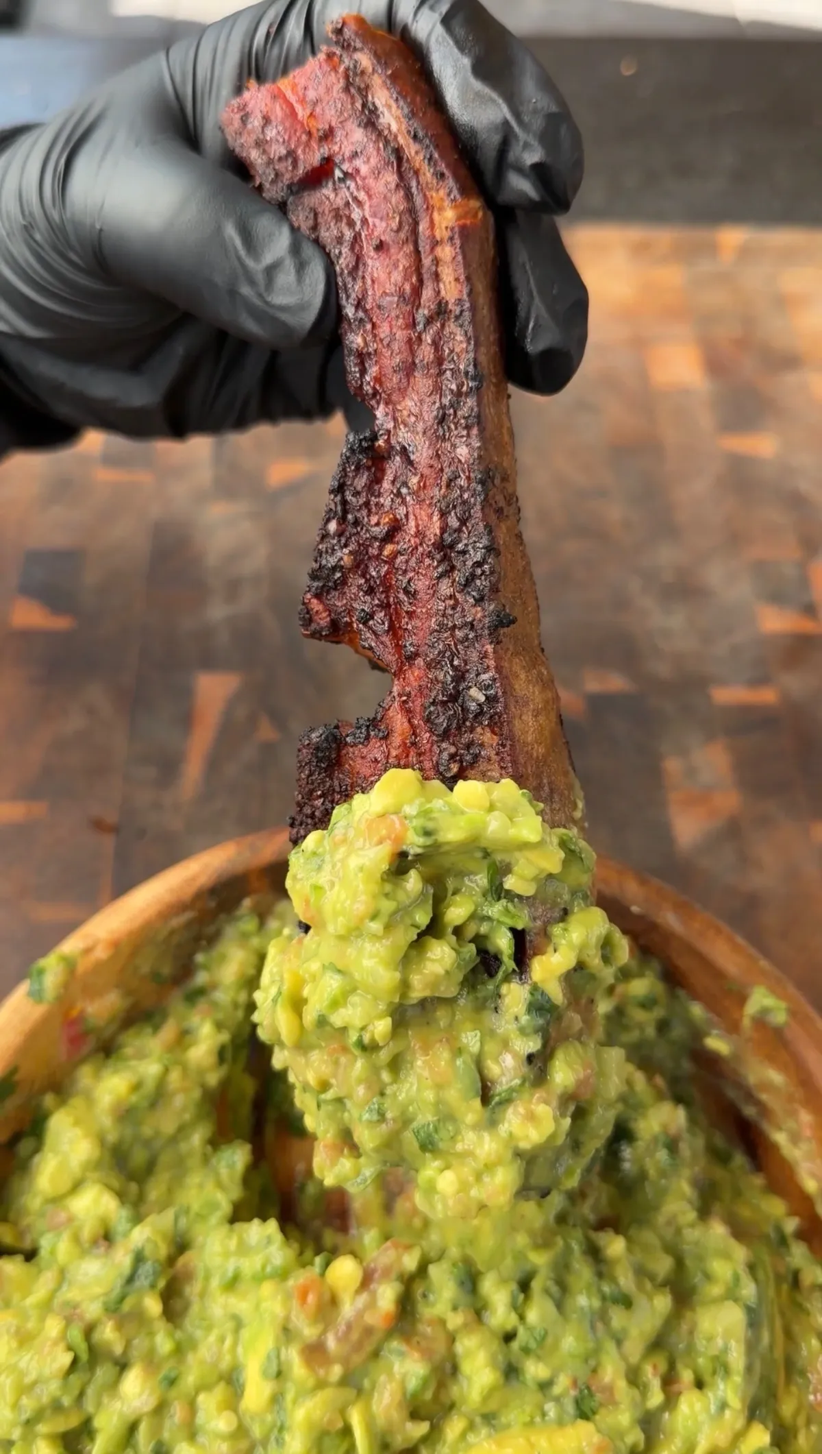 A gloved hand holding a crispy chicharron rib being dipped into a wooden bowl of chunky guacamole