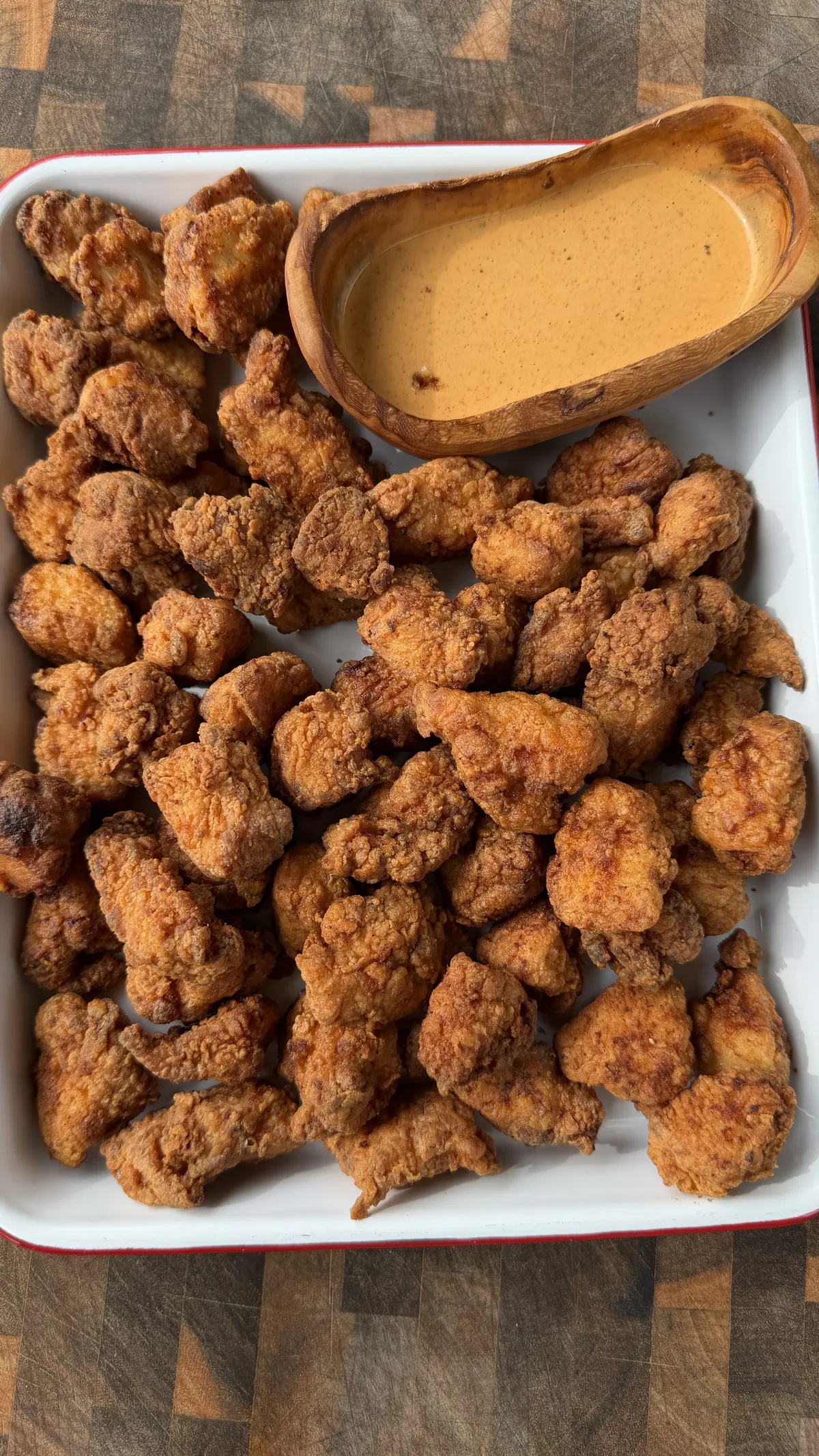 Golden crispy popcorn chicken bites piled on a white tray with a wooden bowl of creamy dipping sauce