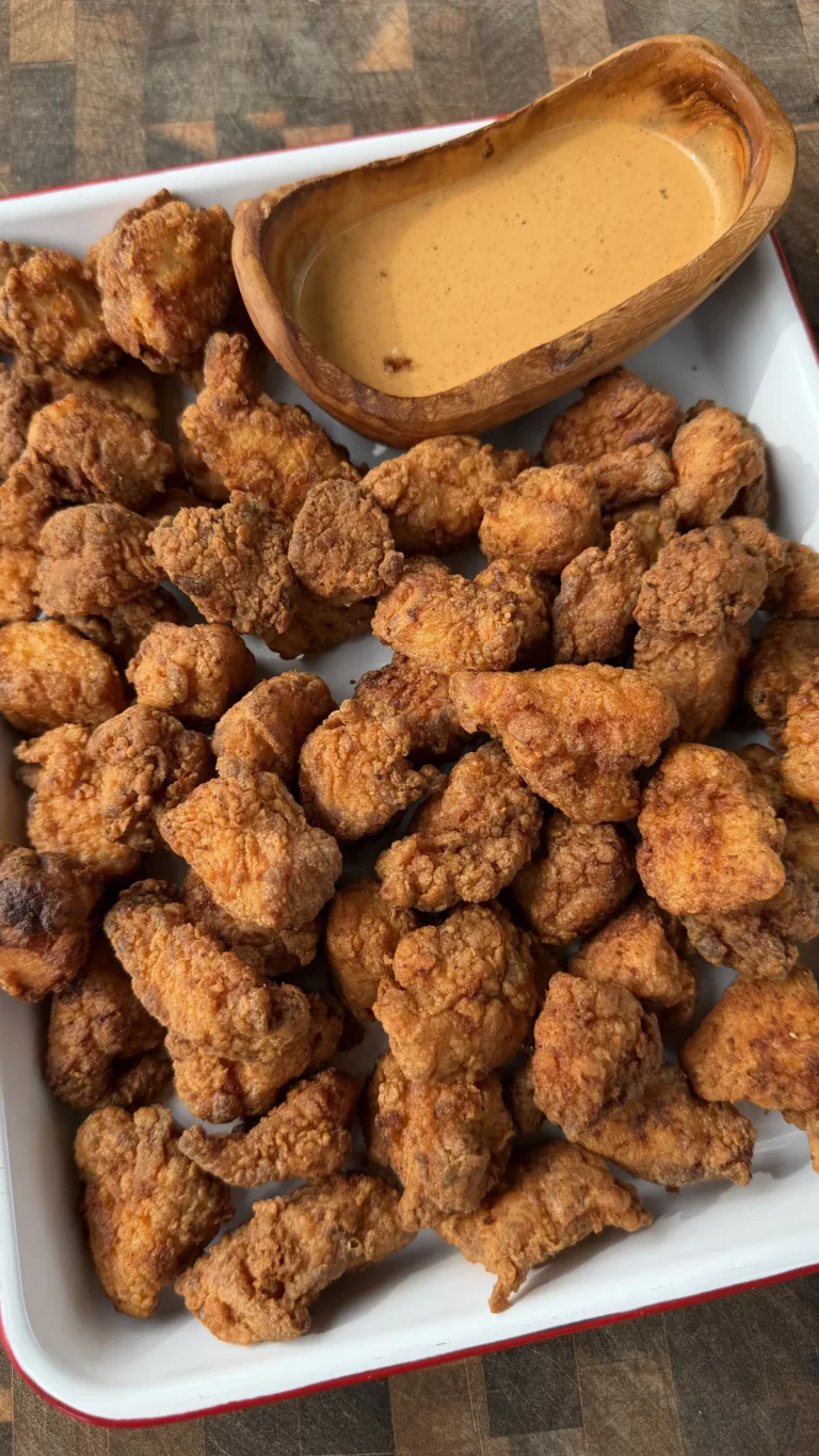 Golden crispy popcorn chicken bites piled on white tray with creamy Cajun dipping sauce in wooden bowl