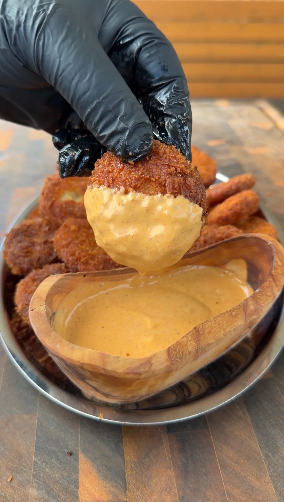 Gloved hand dipping a crispy fried potato cheese ball into creamy orange dipping sauce in a wooden bowl
