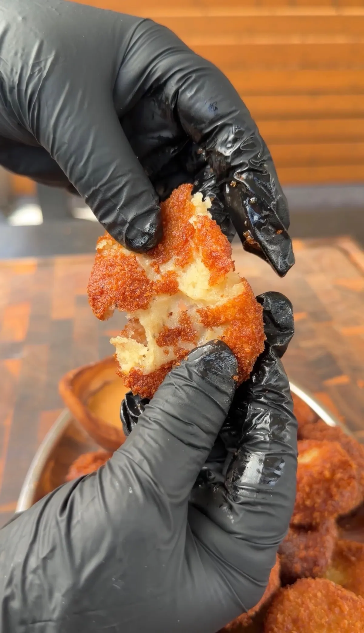 Gloved hands pulling apart a crispy golden potato cheese ball revealing a soft, fluffy interior over a tray of more balls