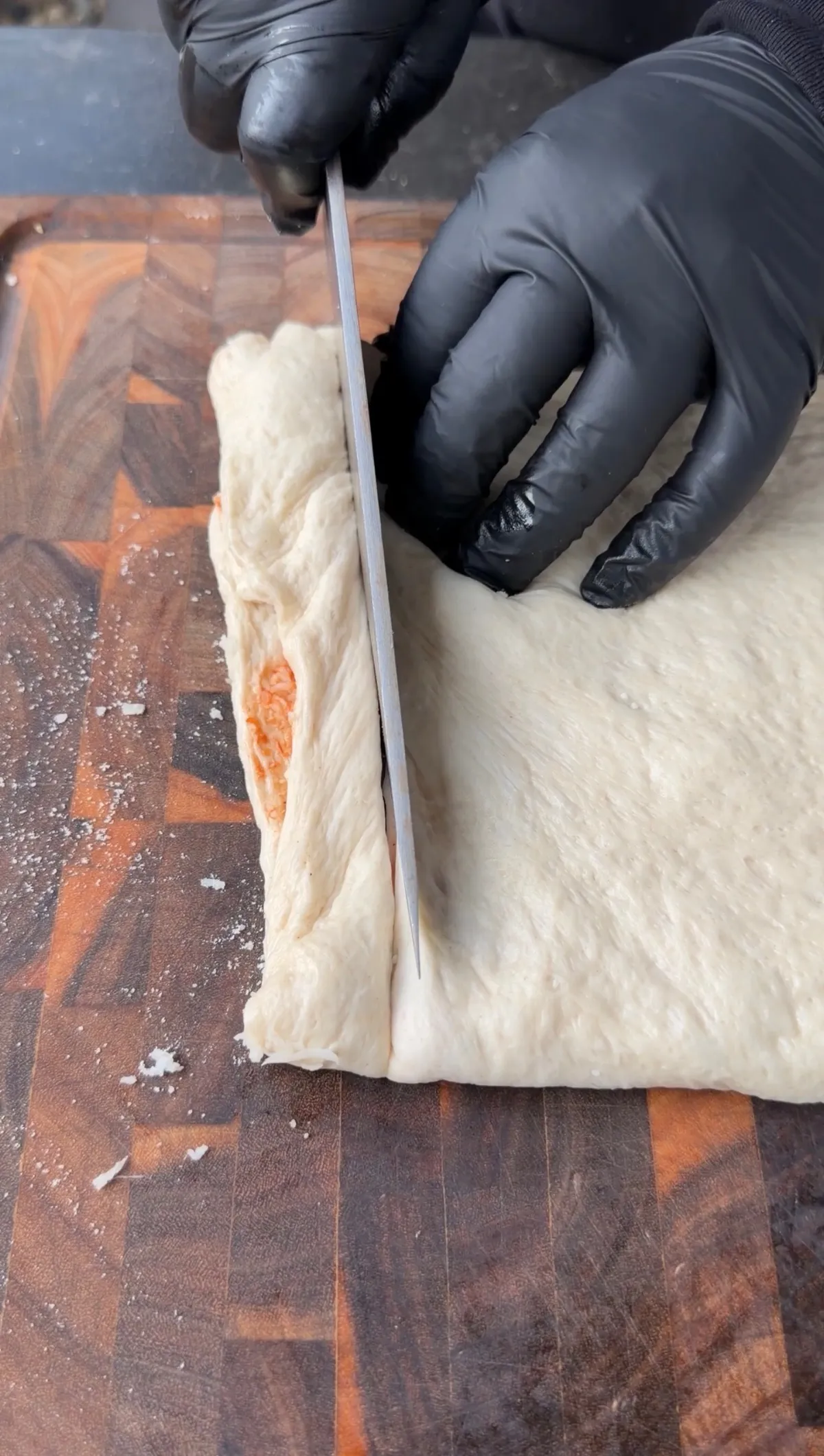 Gloved hands cutting folded Pillsbury pizza dough filled with marinara and mozzarella into strips on a wooden cutting board