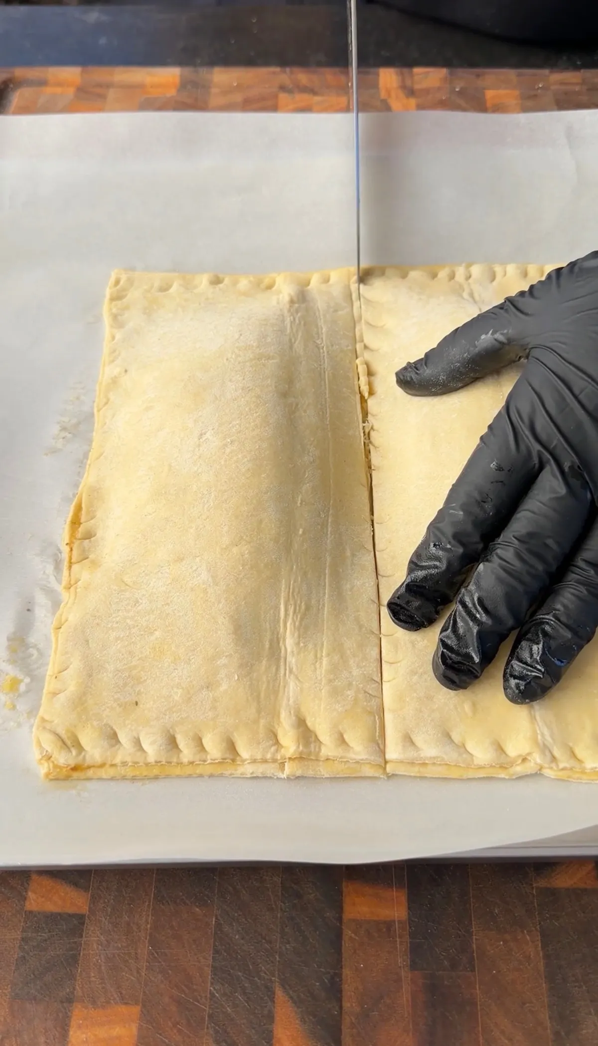 Gloved hand holding unbaked puff pastry cheeseburger hot pockets being sliced in half on parchment paper