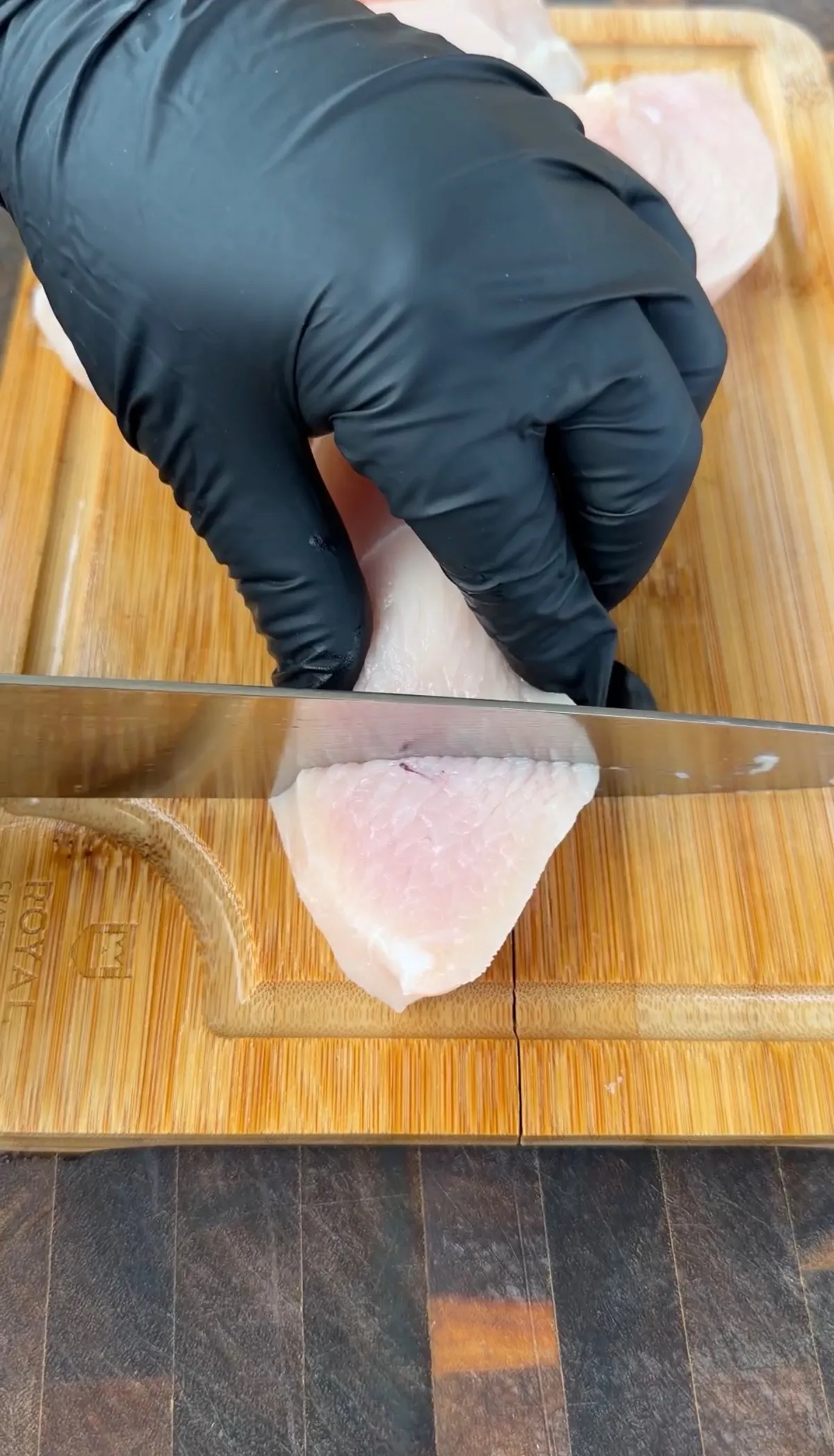 Gloved hand slicing raw chicken breast with a knife on a bamboo cutting board for popcorn chicken