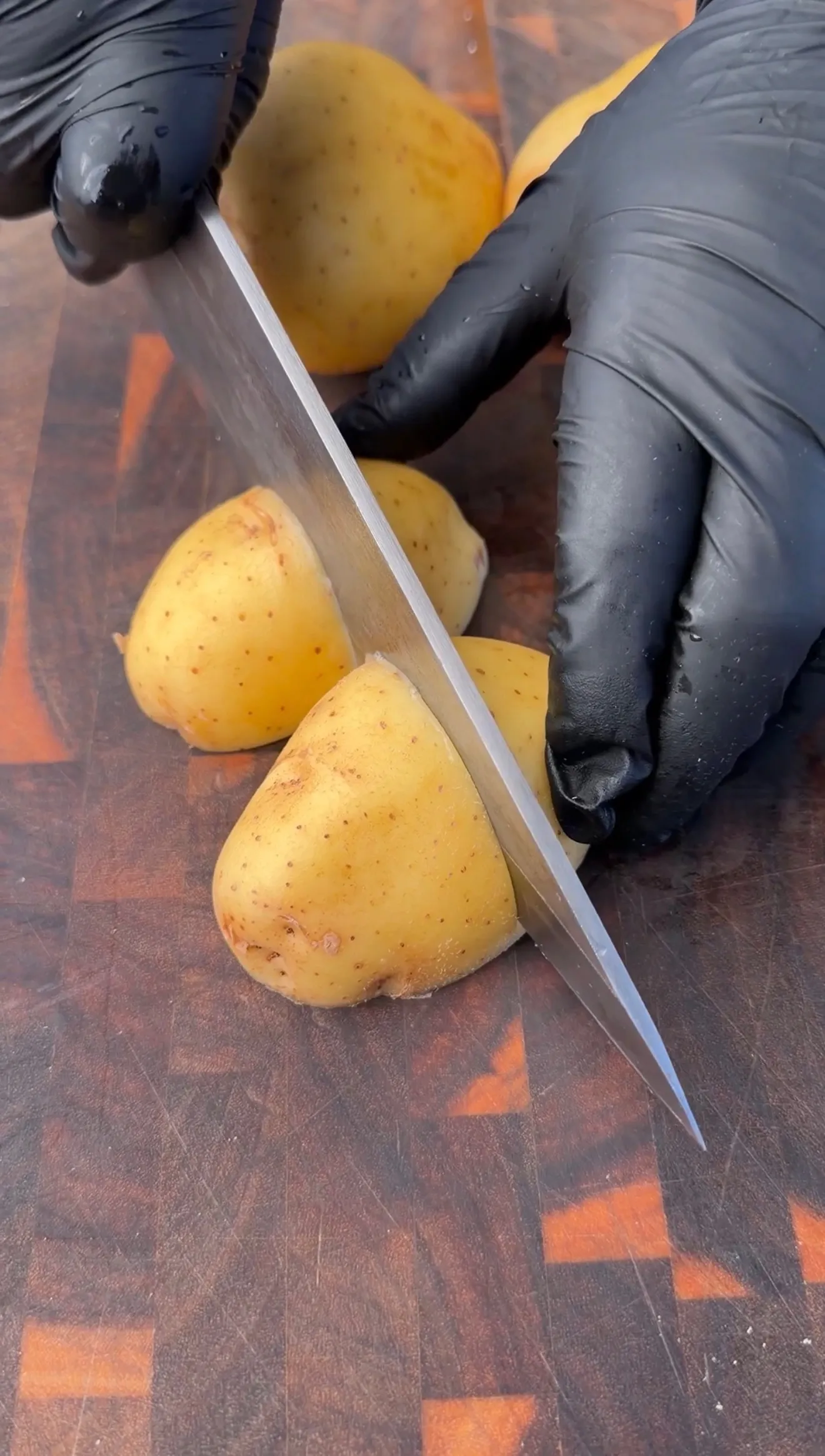 Hands in black gloves cutting fresh Yukon Gold potatoes on a wooden cutting board with a chef's knife