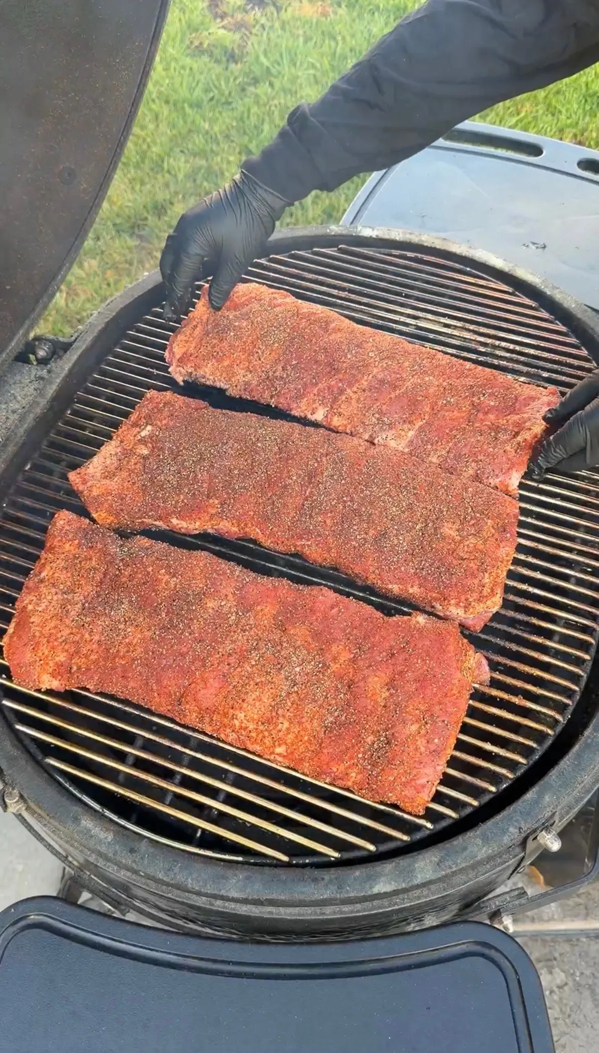 Three racks of dry-rubbed pork ribs being placed on a round charcoal grill by hands in black gloves