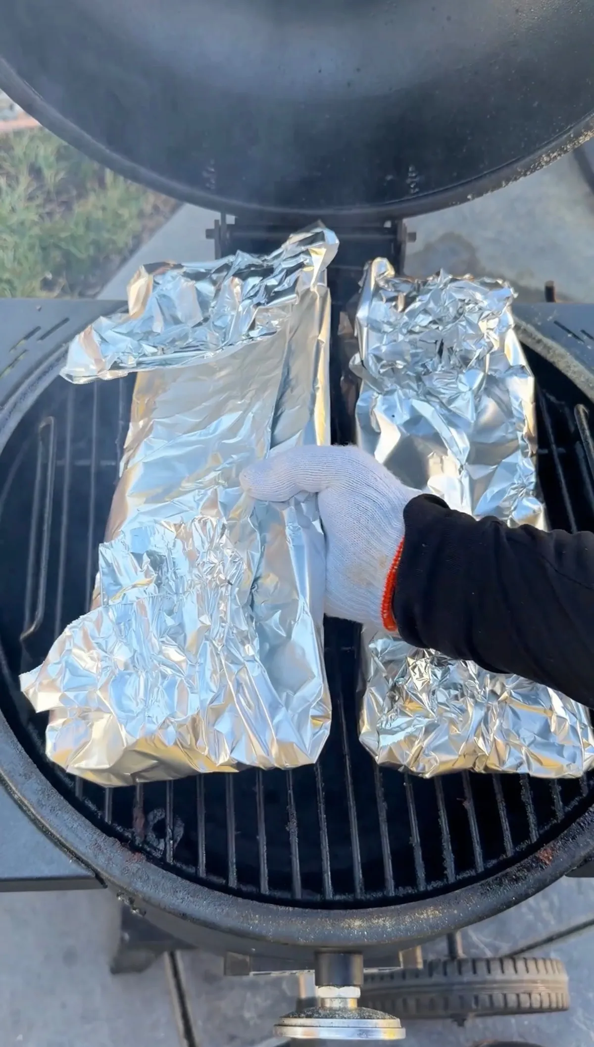 Foil-wrapped St. Louis spare ribs arranged on a round charcoal smoker grill, handled with heat-resistant gloves