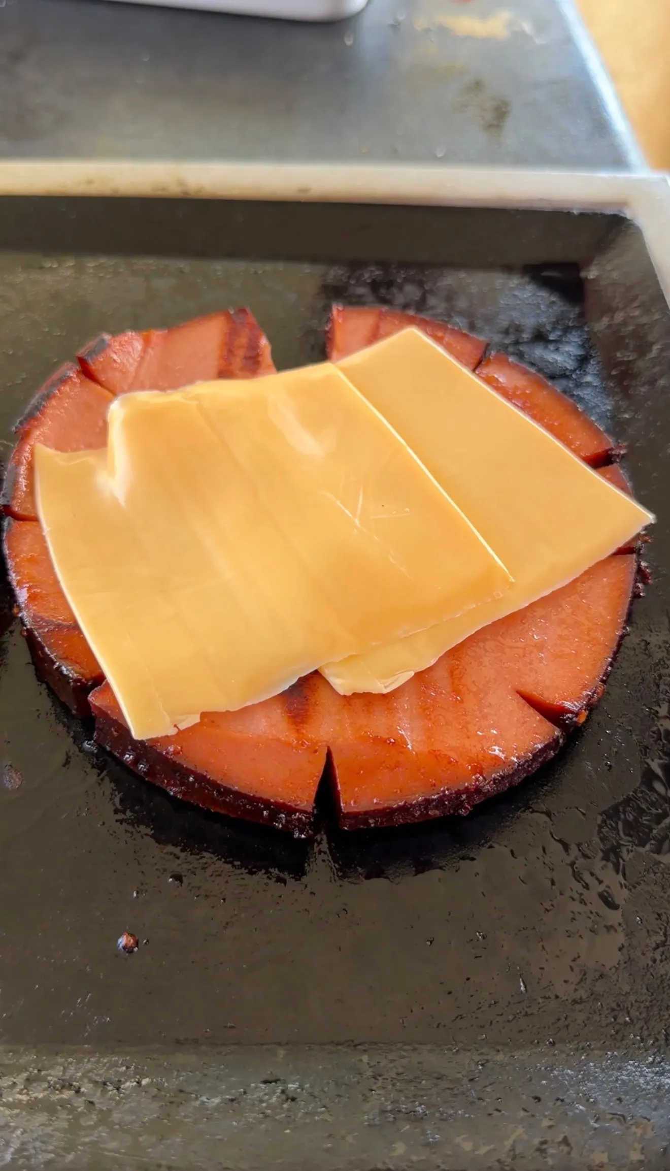 Thick-cut smoked bologna slice with cuts around the edges topped with American cheese melting on a flat top griddle