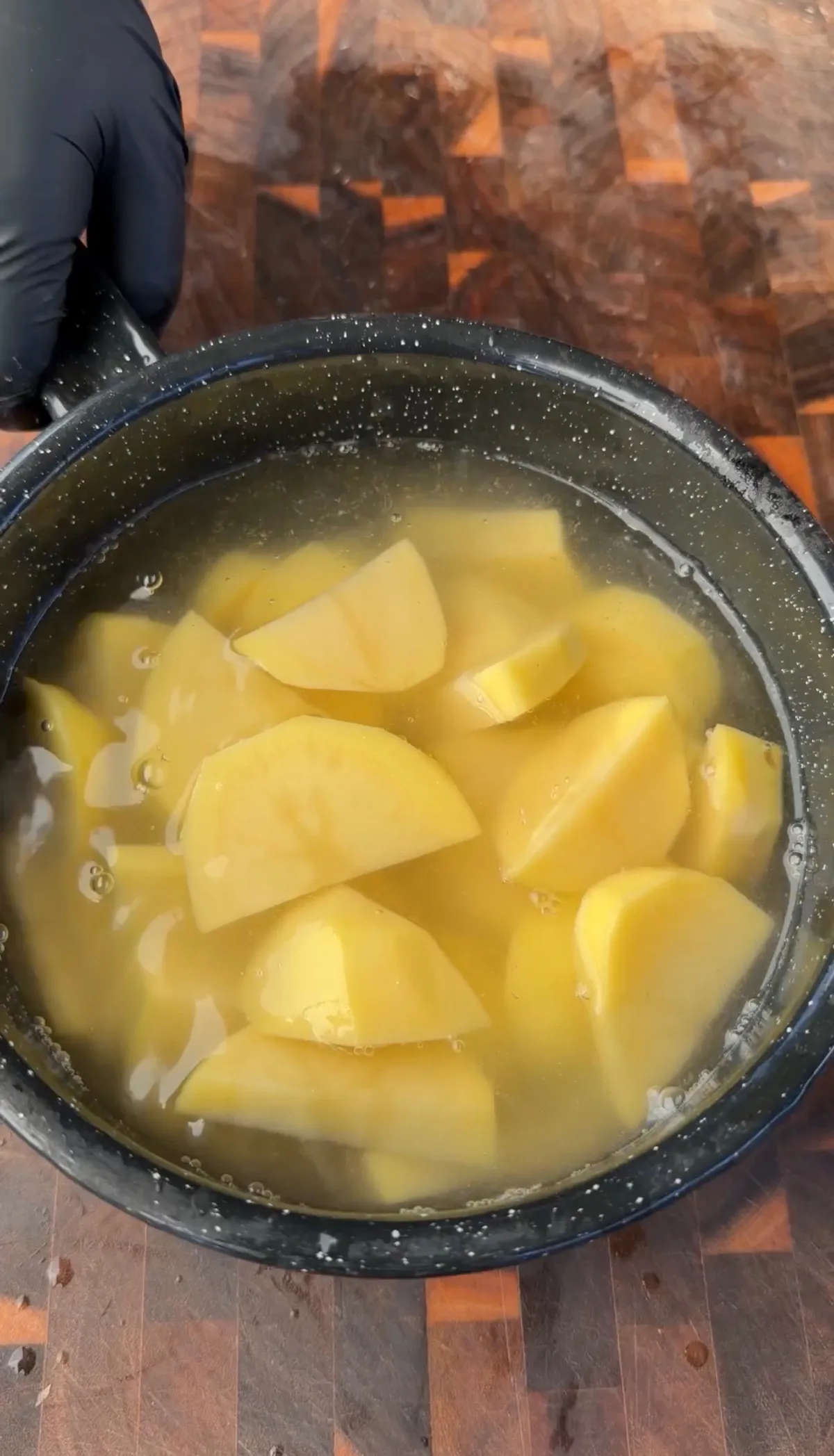 Peeled golden potato chunks boiling in salted water in a dark pot on a wooden cutting board