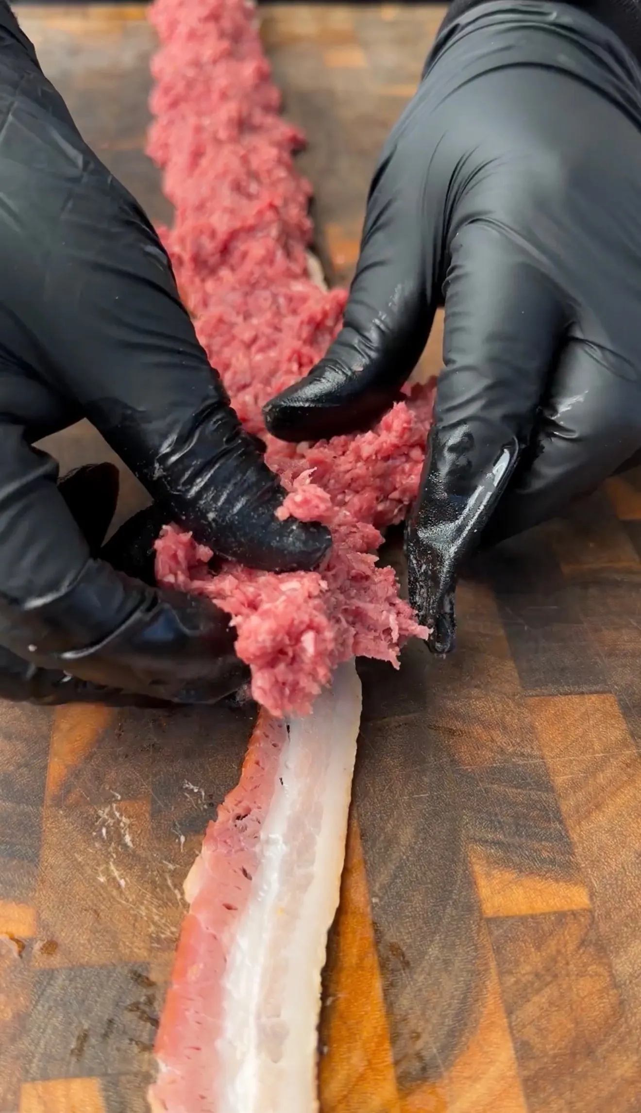 Hands in black gloves spreading seasoned ground beef mixture onto raw bacon strips on a wooden cutting board