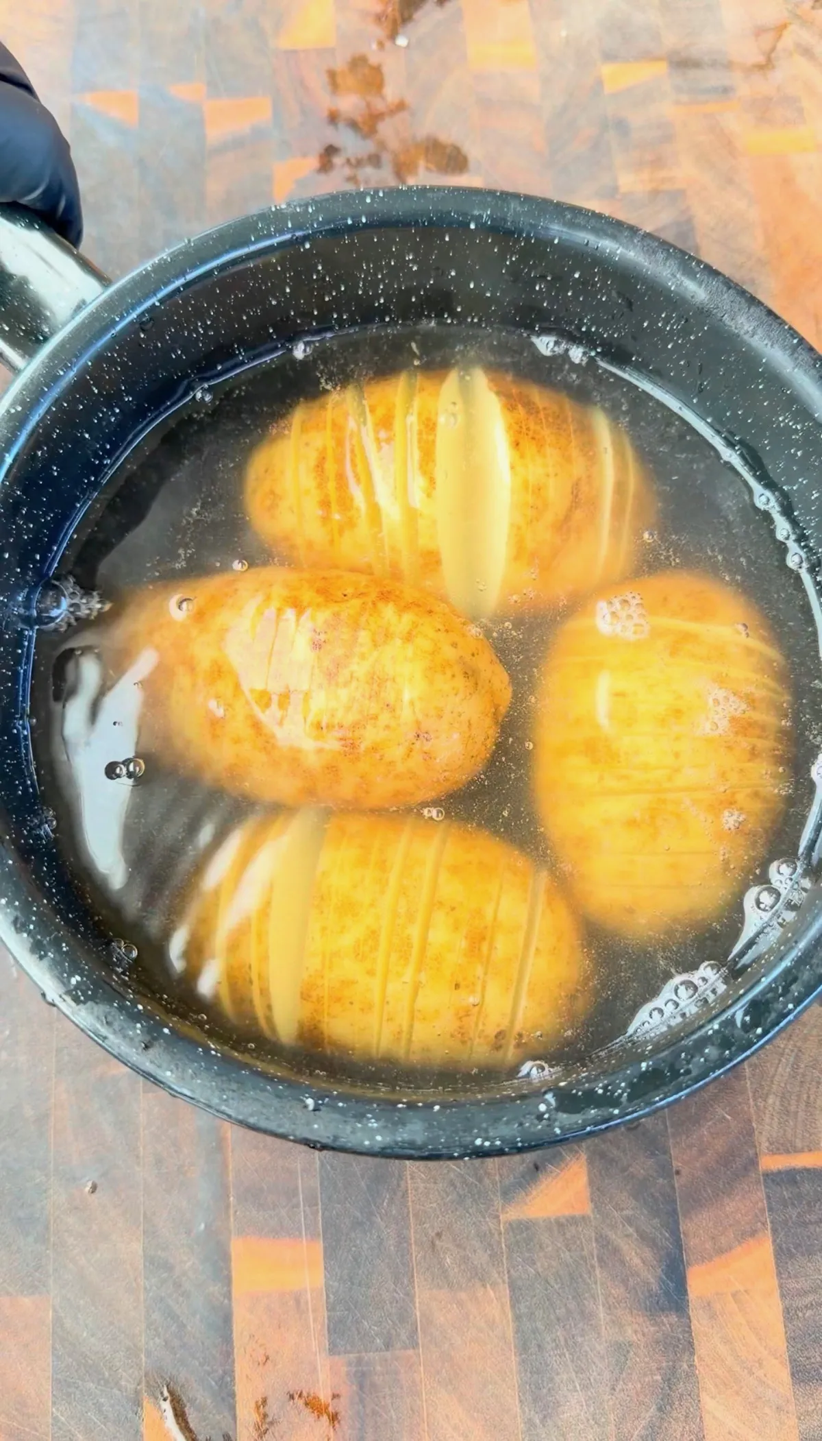 Four seasoned hasselback potatoes with accordion cuts simmering in a dark skillet on a wood cutting board