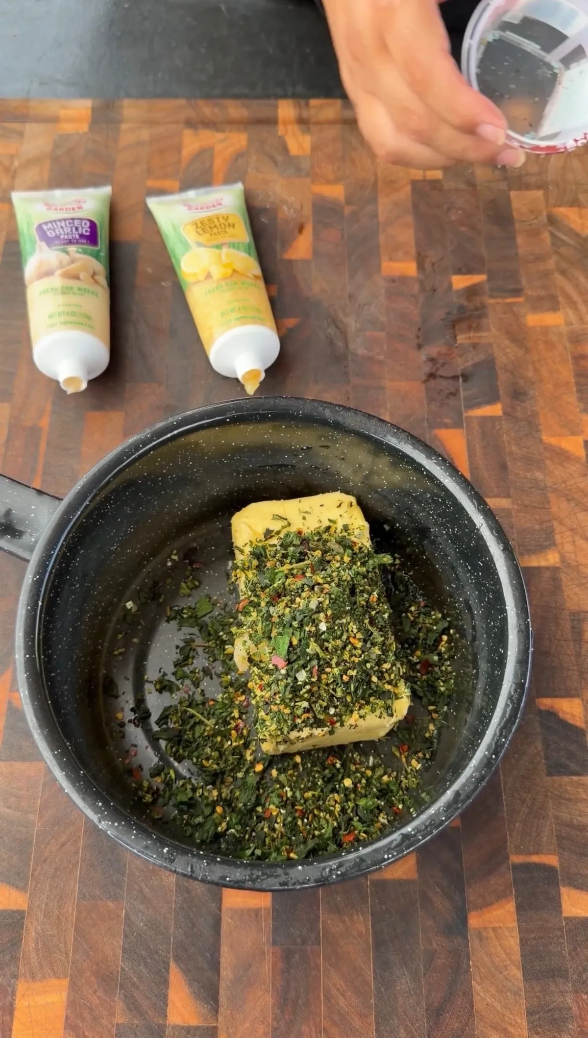 Stick of butter topped with mixed herbs in a black saucepan with minced garlic and zesty lemon paste tubes nearby
