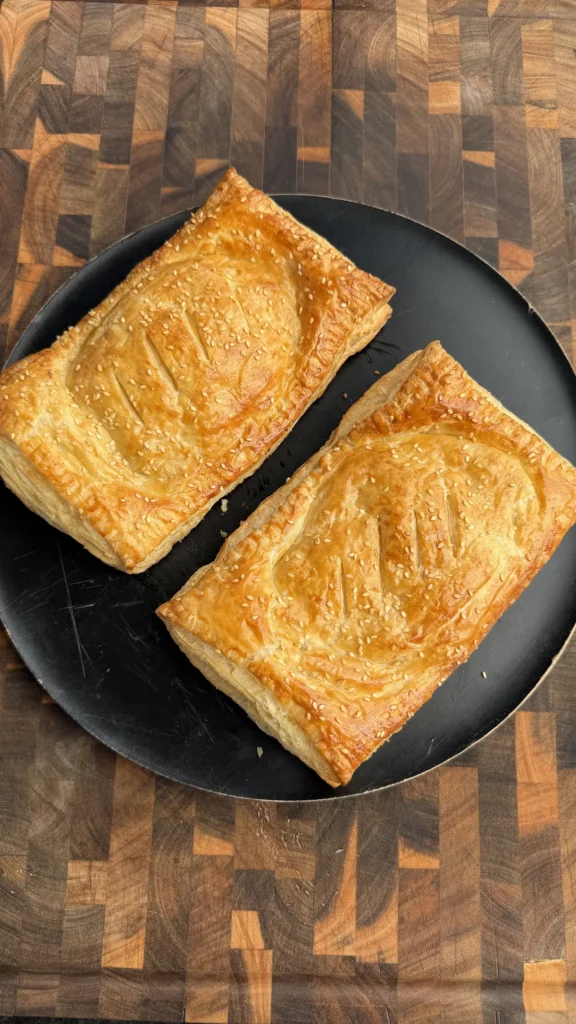 Two golden baked cheeseburger hot pockets made with puff pastry, topped with sesame seeds, on a black plate