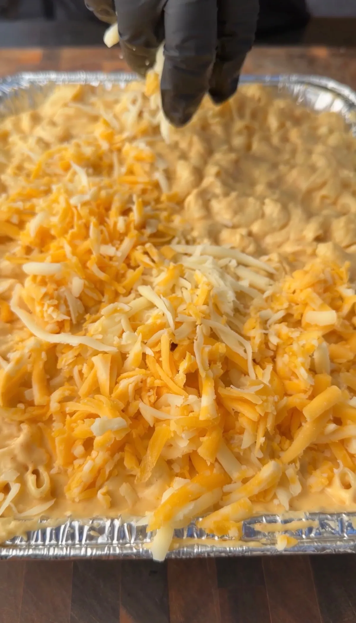 Gloved hand sprinkling freshly shredded Colby Jack and sharp cheddar over elbow macaroni and cheese in a foil baking dish