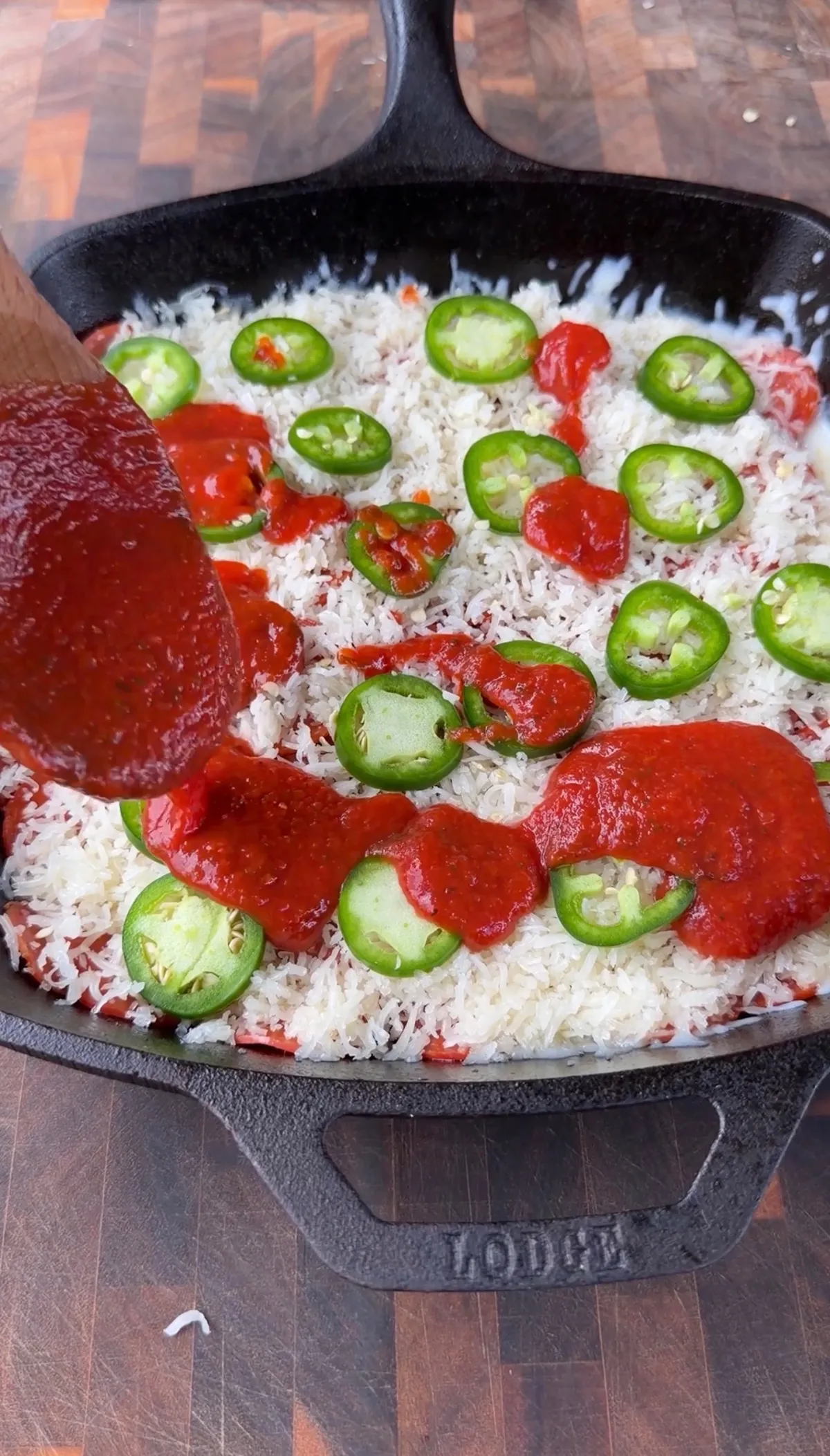 Marinara sauce being spooned over shredded mozzarella and sliced fresh jalapeños in a Lodge cast-iron skillet