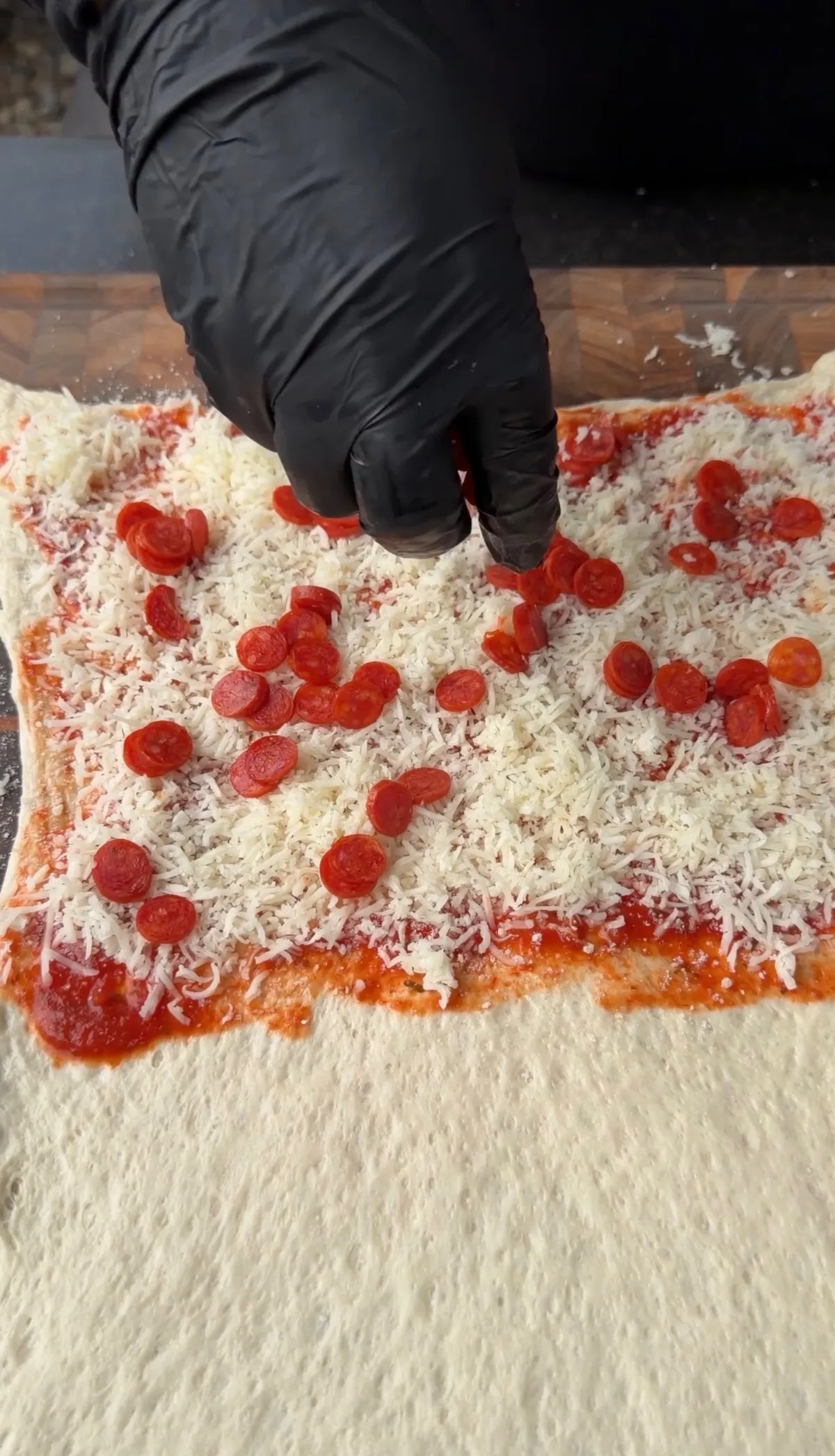 Gloved hand sprinkling mini pepperonis over freshly grated mozzarella and marinara sauce on rolled pizza dough