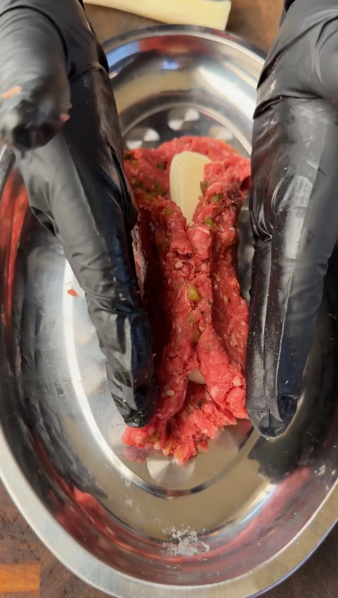 Gloved hands mixing seasoned ground beef with herbs and garlic in a stainless steel bowl for cheese stuffed burger dogs
