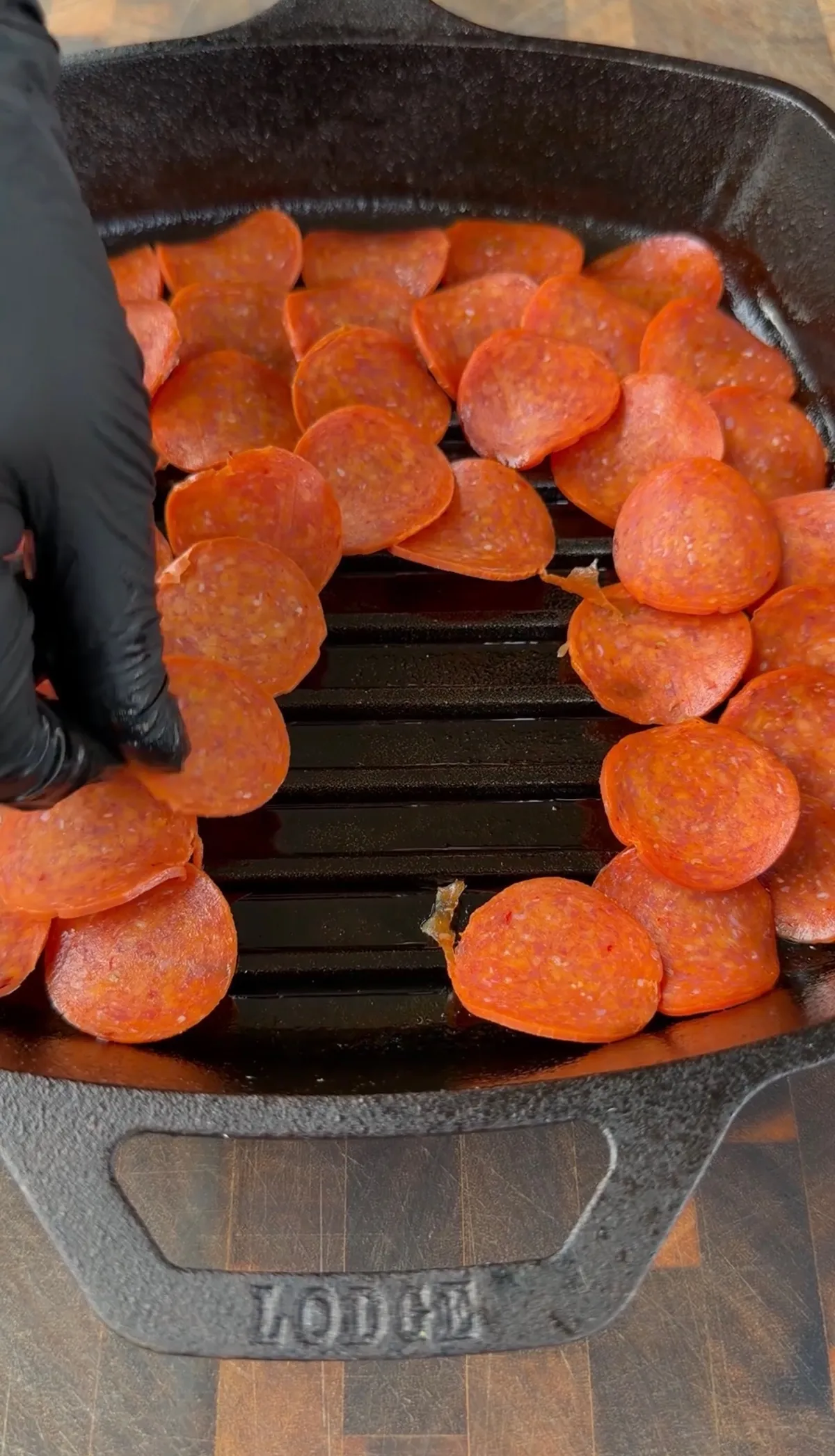 Gloved hand laying overlapping regular-size pepperoni slices across a Lodge cast-iron grill pan for upside-down deep dish pizza