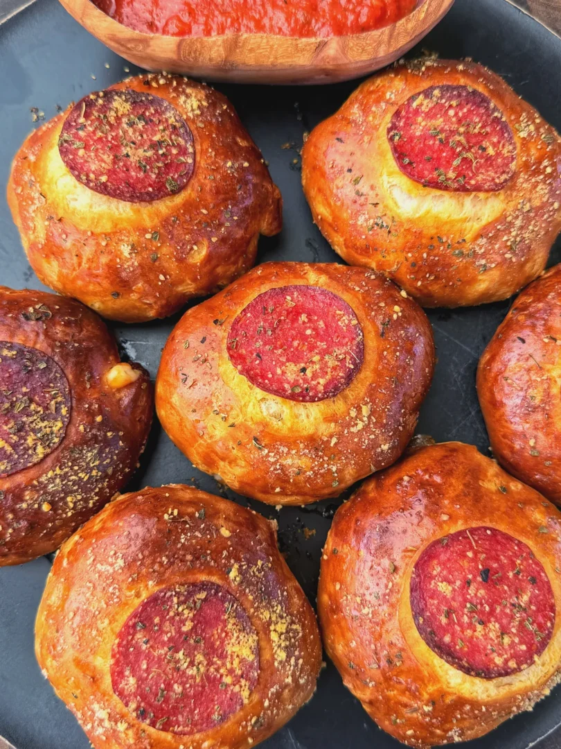 Golden baked pepperoni pizza bombs topped with Italian seasoning on a dark plate with marinara dipping sauce
