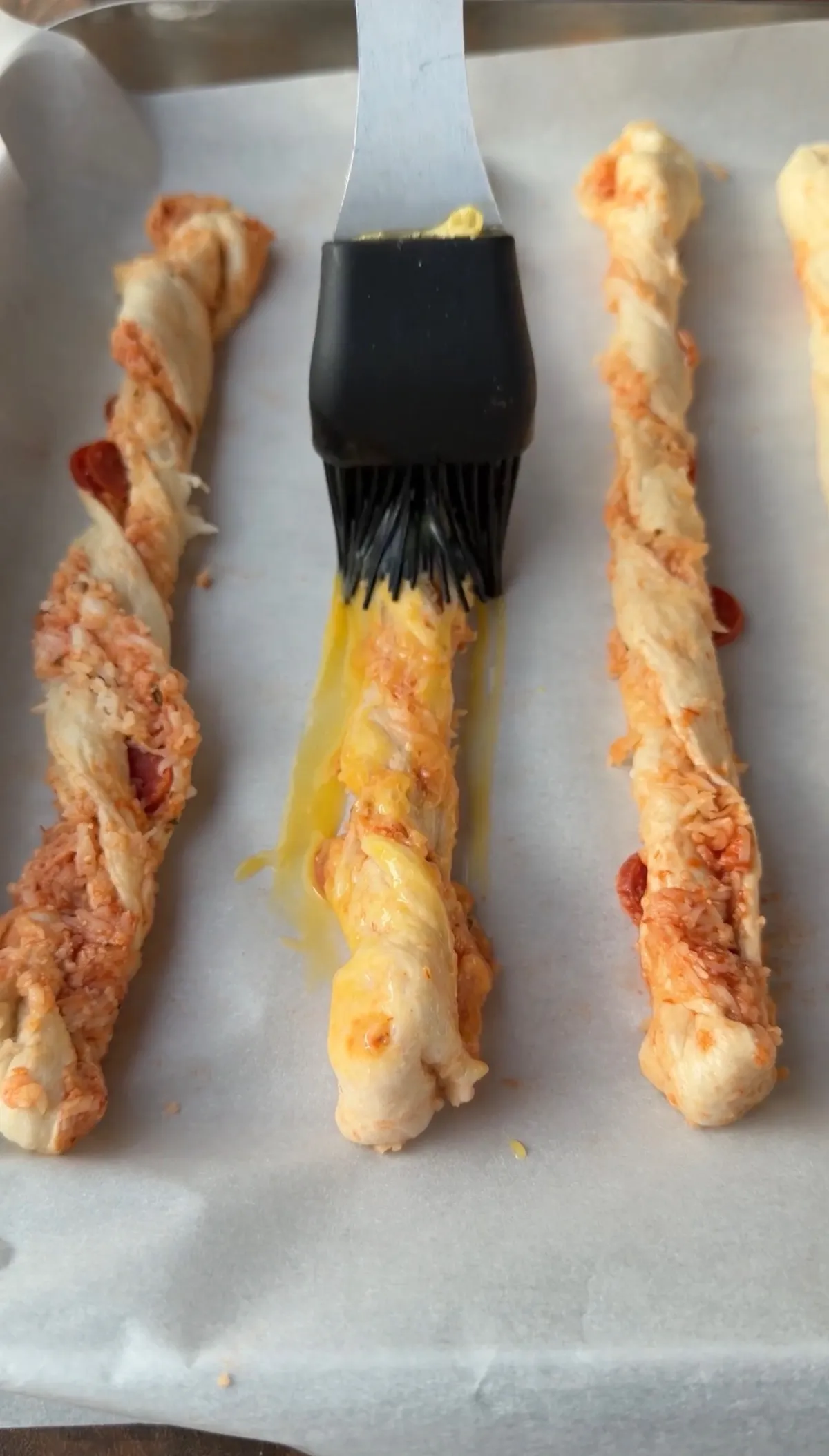 Brushing egg wash onto twisted pepperoni pizza twists with mini pepperonis on a parchment-lined baking sheet