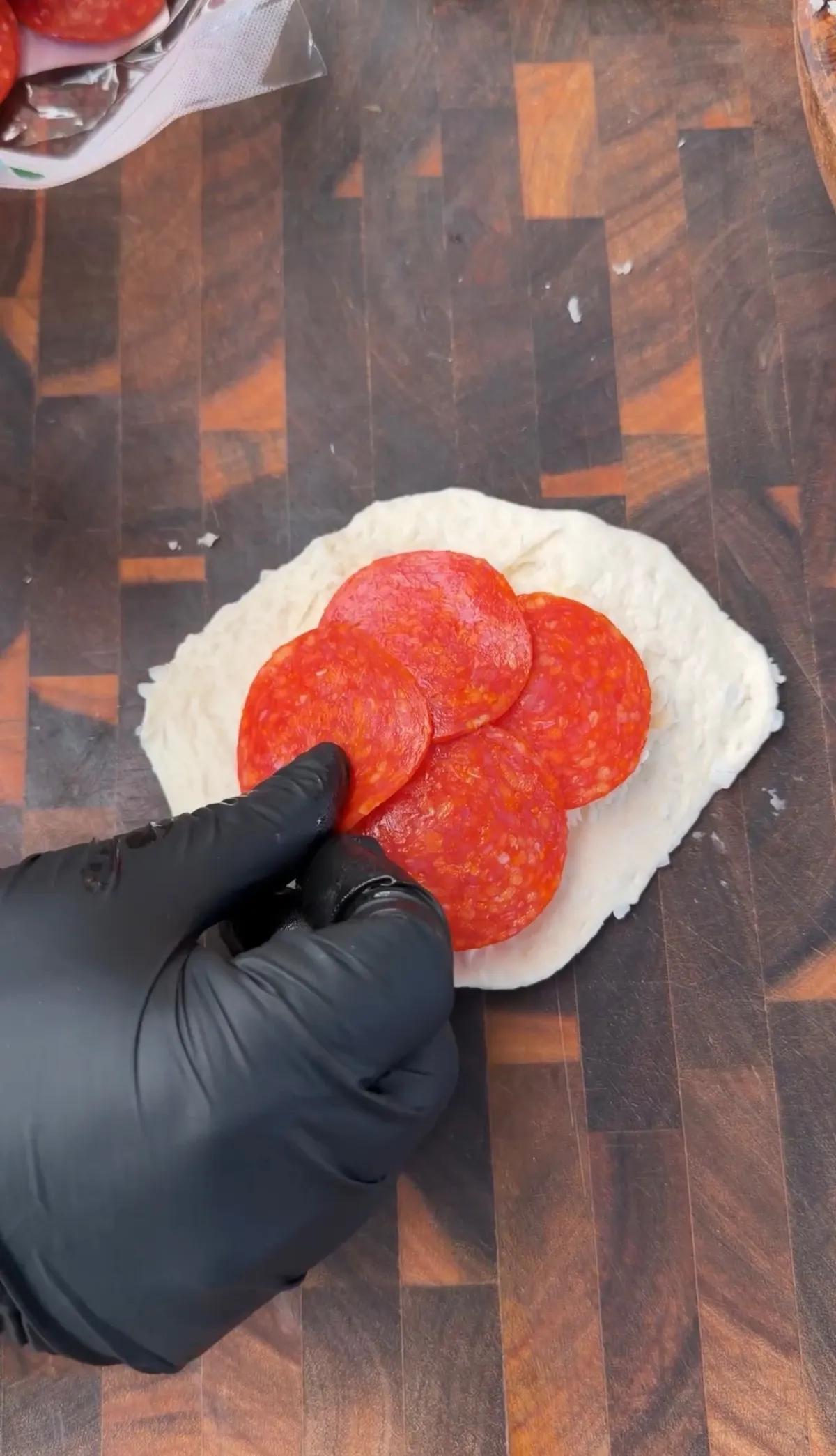 Gloved hand placing pepperoni slices onto flattened pizza dough on a wooden cutting board for pizza bombs