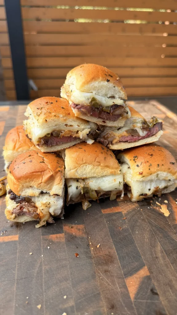 Stacked picanha steak sliders on toasted King's Hawaiian rolls with melted provolone, sautéed peppers and onions on a wood cutting board