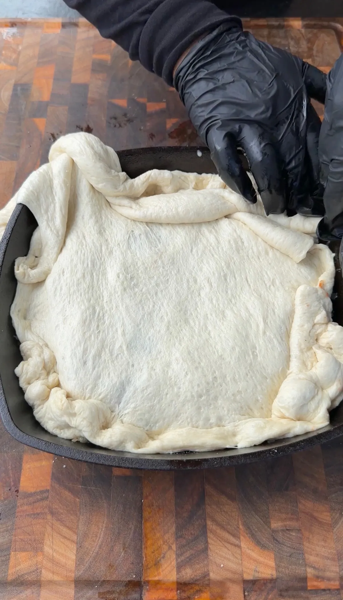 Hands in black gloves tucking Pillsbury pizza dough over upside-down deep dish jalapeño pepperoni pizza in cast-iron skillet
