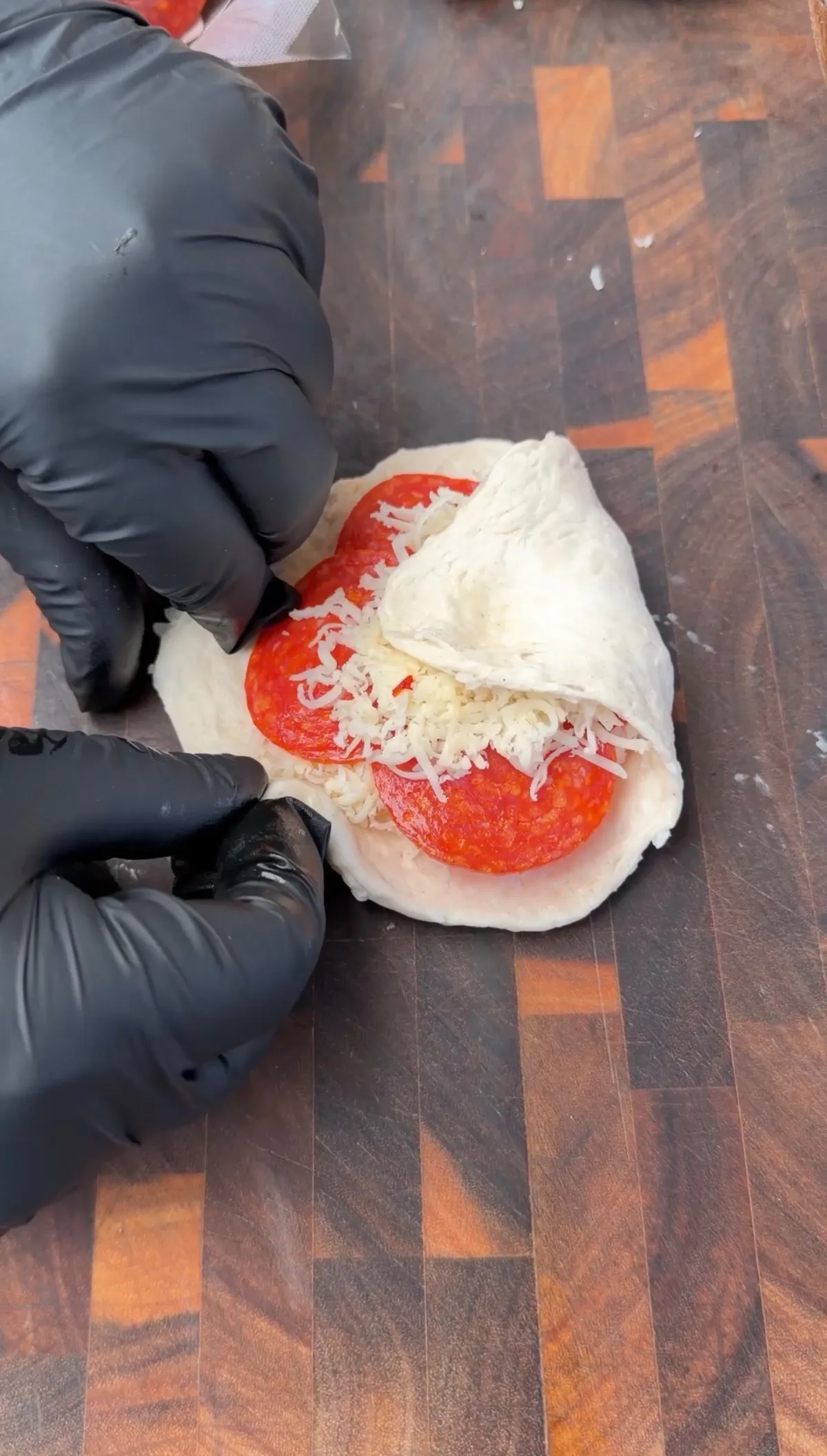Hands in black gloves folding pizza dough over pepperoni and shredded mozzarella on a wooden cutting board