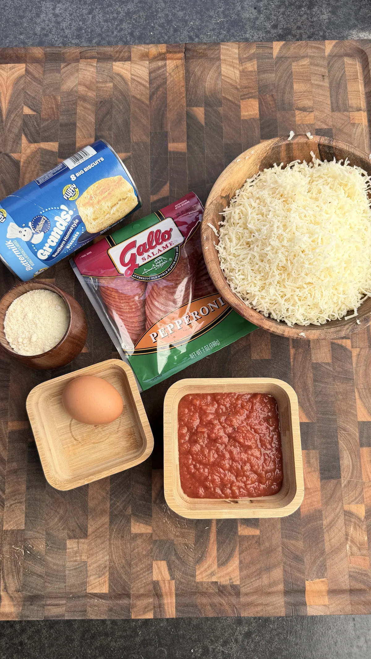 Pizza bomb ingredients: Grands biscuits, Gallo pepperoni, shredded mozzarella, marinara sauce, egg, and breadcrumbs on wooden board
