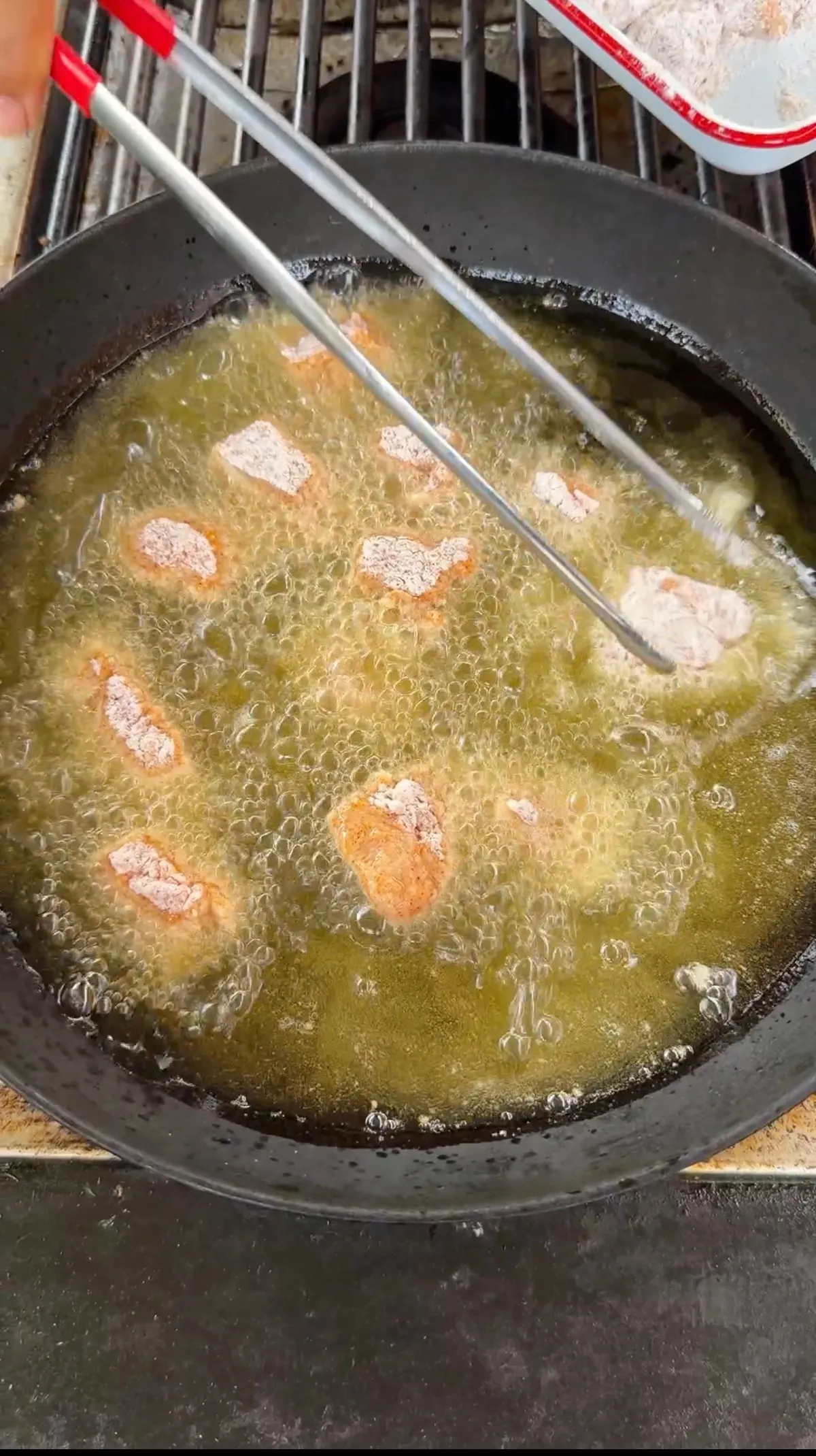 Bite-sized popcorn chicken pieces deep frying in bubbling hot oil in a cast iron skillet with tongs