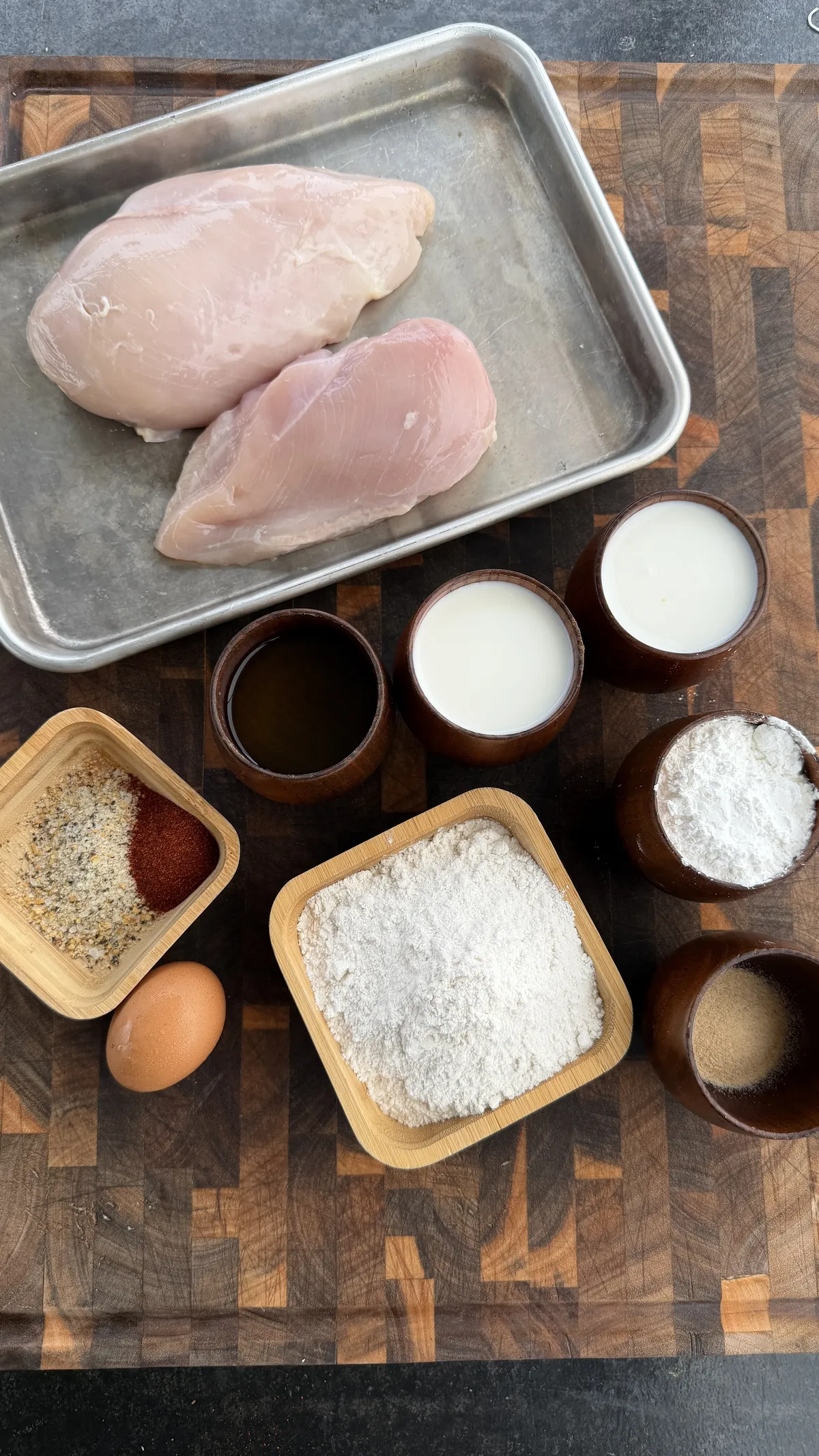 Raw chicken breasts with flour, egg, milk, spices, and cornstarch ingredients for popcorn chicken on a wooden board